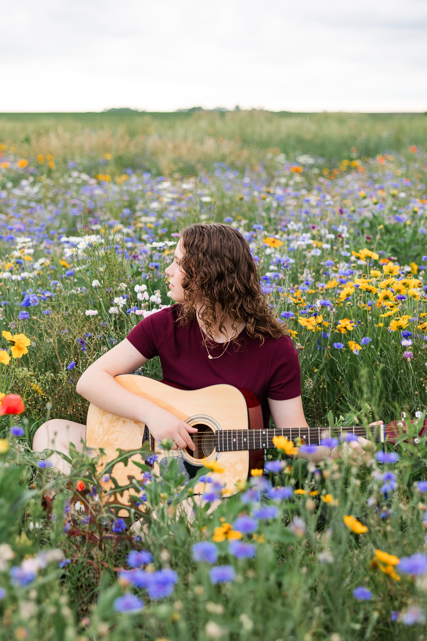 Alaina sits and looks off into the distance while she plays her guitar in a colorful wildflower field in Iowa | CB Studio