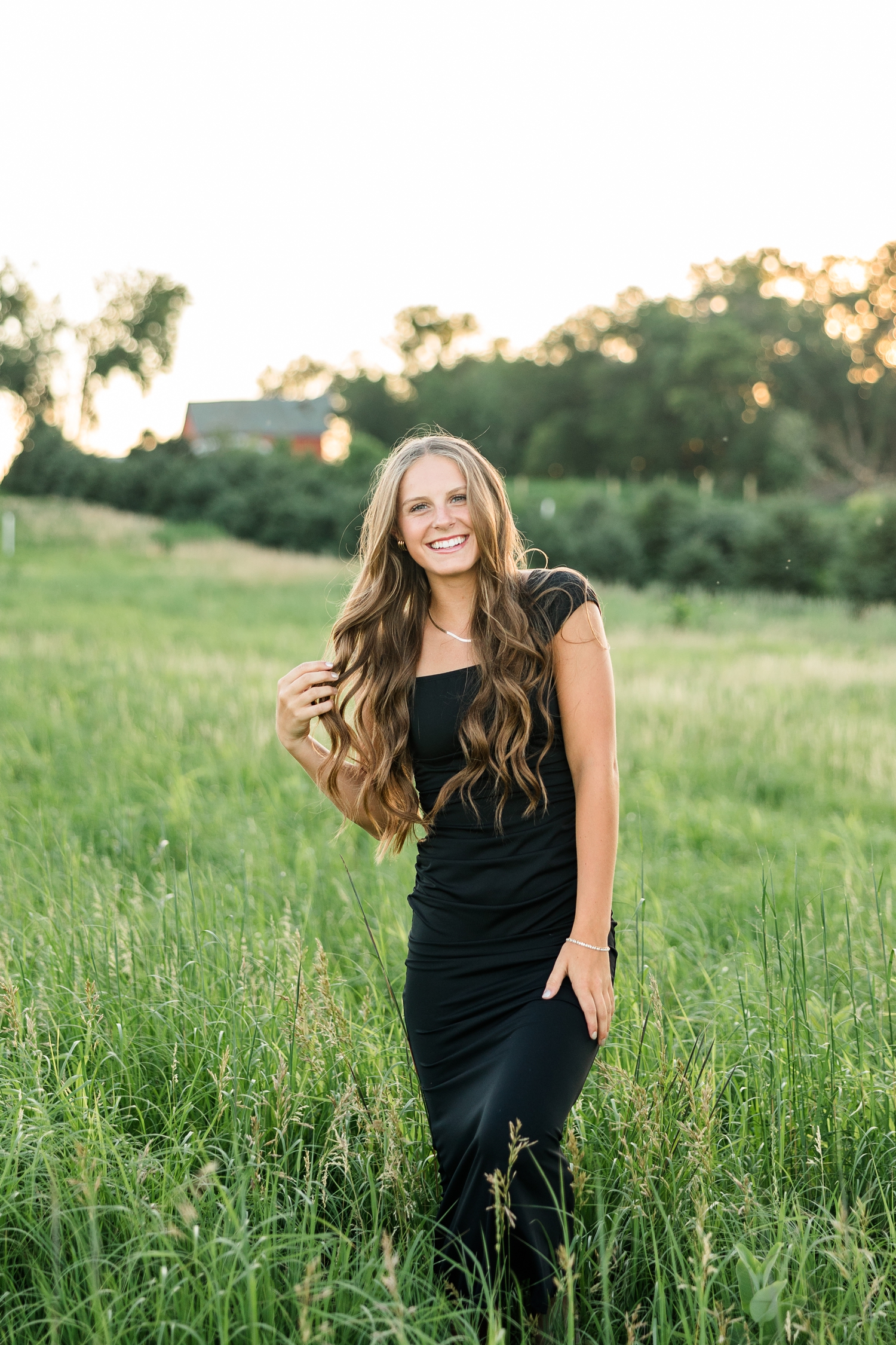 Ashton, wearing a black formal dress, laughs as she walks in a grassy pasture with a barn in the background | CB Studio