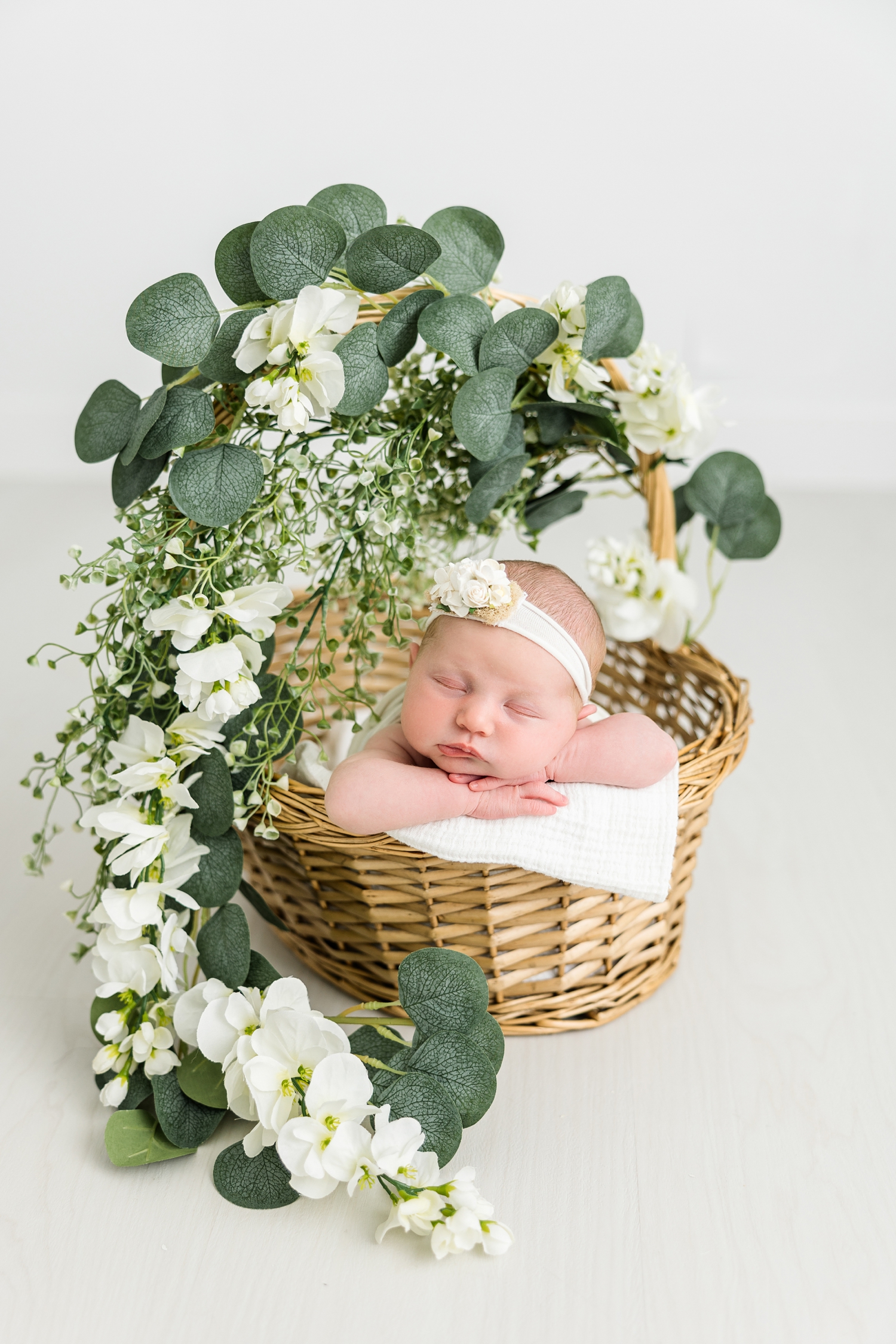 In an all white room, baby Lylah lays in a wicker basket with greenery and white flowers draped along the the handle | CB Studio