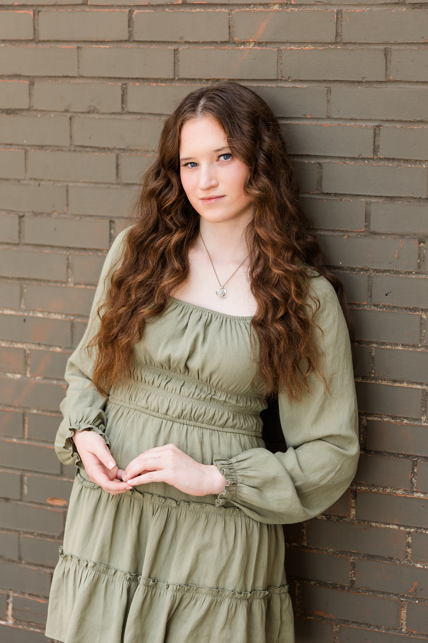 Evie, wearing a sage green dress, leans against an olive green brick wall in downtown Algona, IA| CB Studio