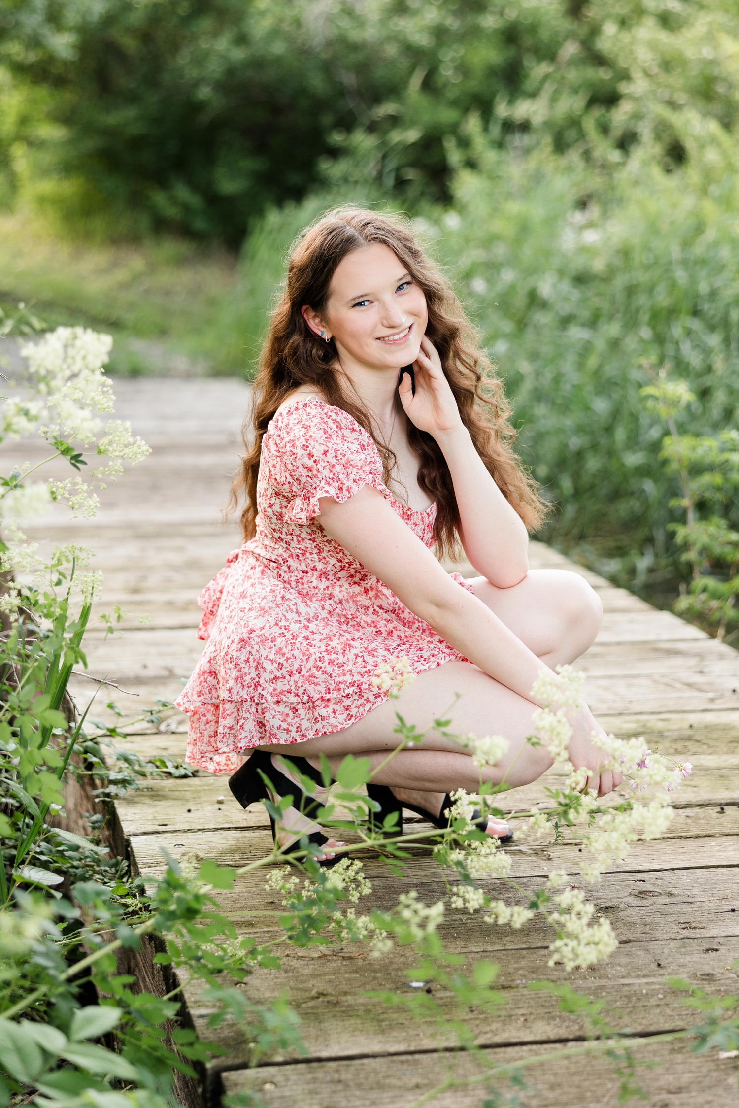 Evie, wearing a pink floral romper, squats in the middle of a wooden bridge path at Water's Edge Nature Center with her hand on her cheek | CB Studio