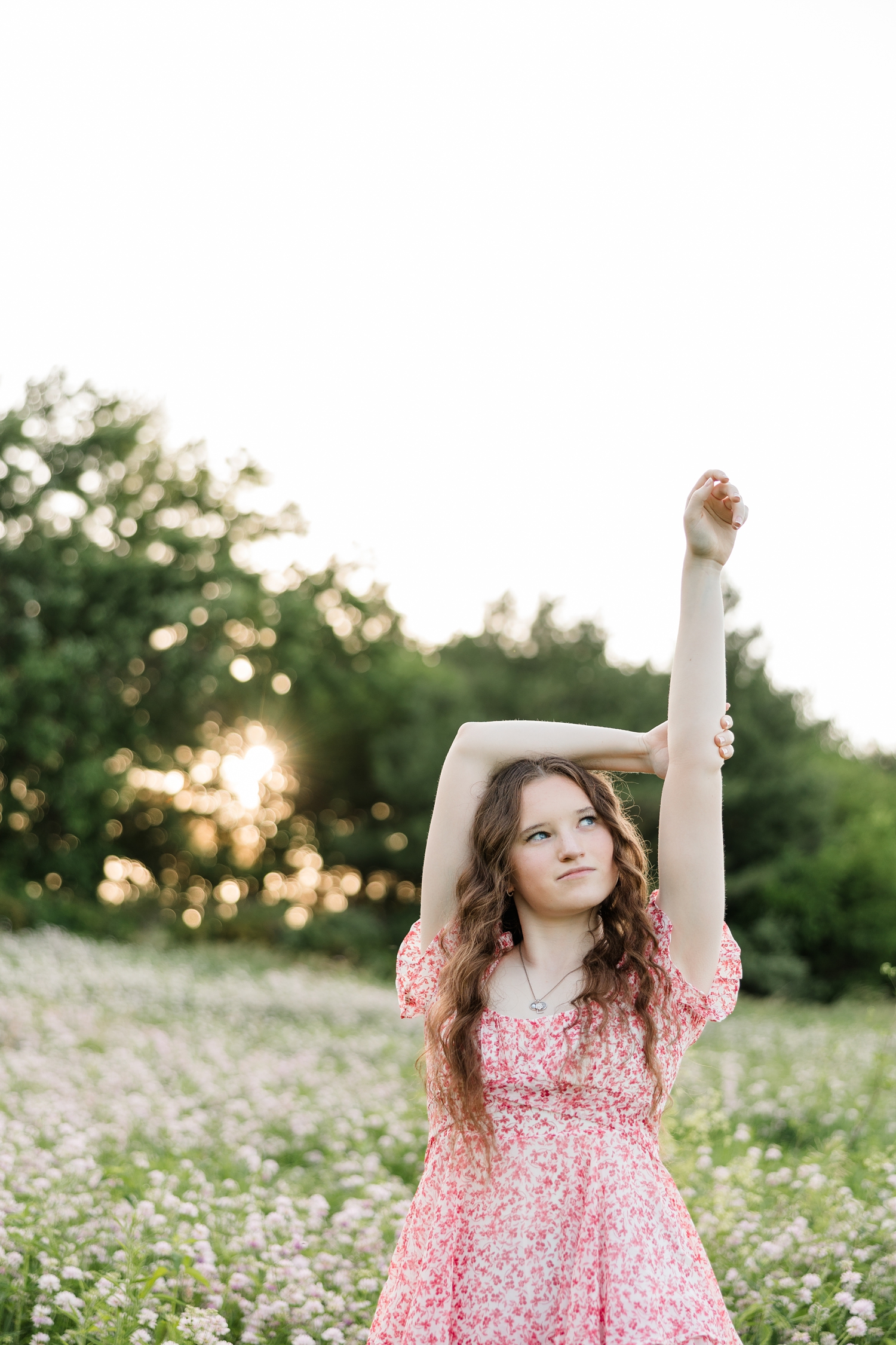 Evie, wearing a pink floral romper, stands in the middle of a flower field at Water's Edge Nature Center with her hands and arms in the air as the sun sets behind the line of trees behind her | CB Studio