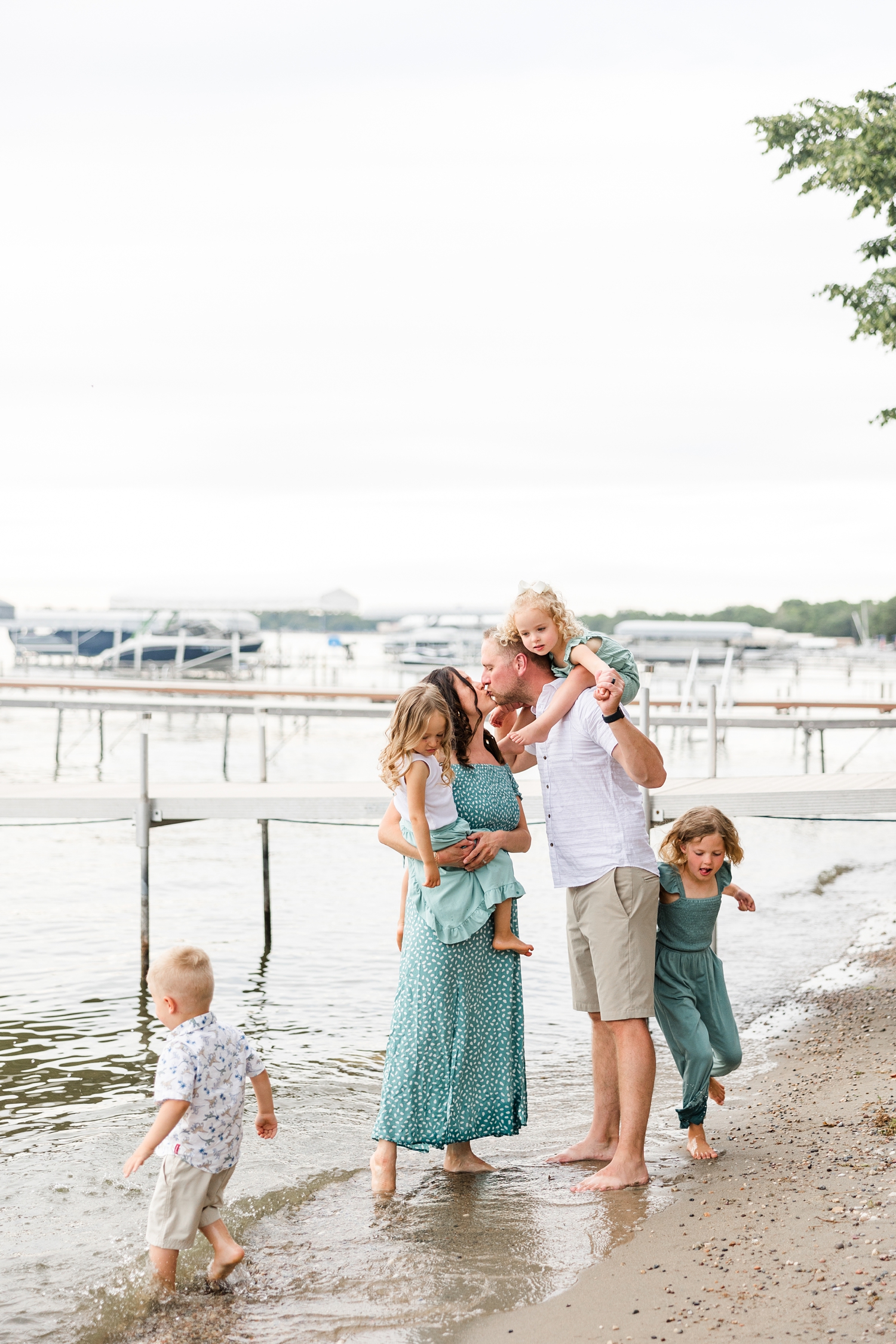 Mom and dad of triplets plus one hold two toddlers as they kiss on the shore of Clear Lake, IA while two other children run around them | CB Studio