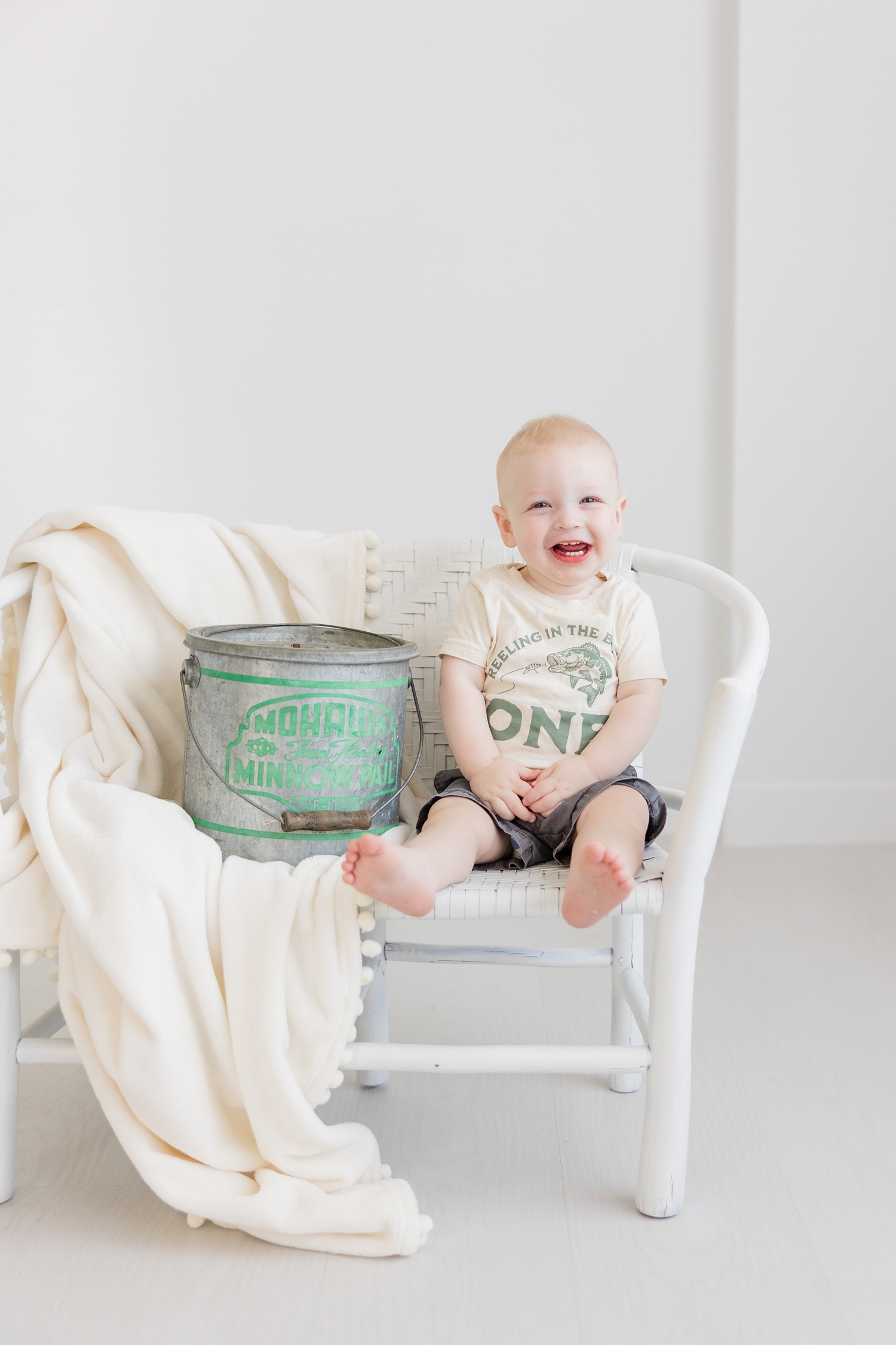 Baby Jonah, wearing a shirt that says "Reeling in the Big ONE," sits on a white wicker bench with a giant smile and a metal, vintage fishing pail with a green logo imprinted | CB Studio