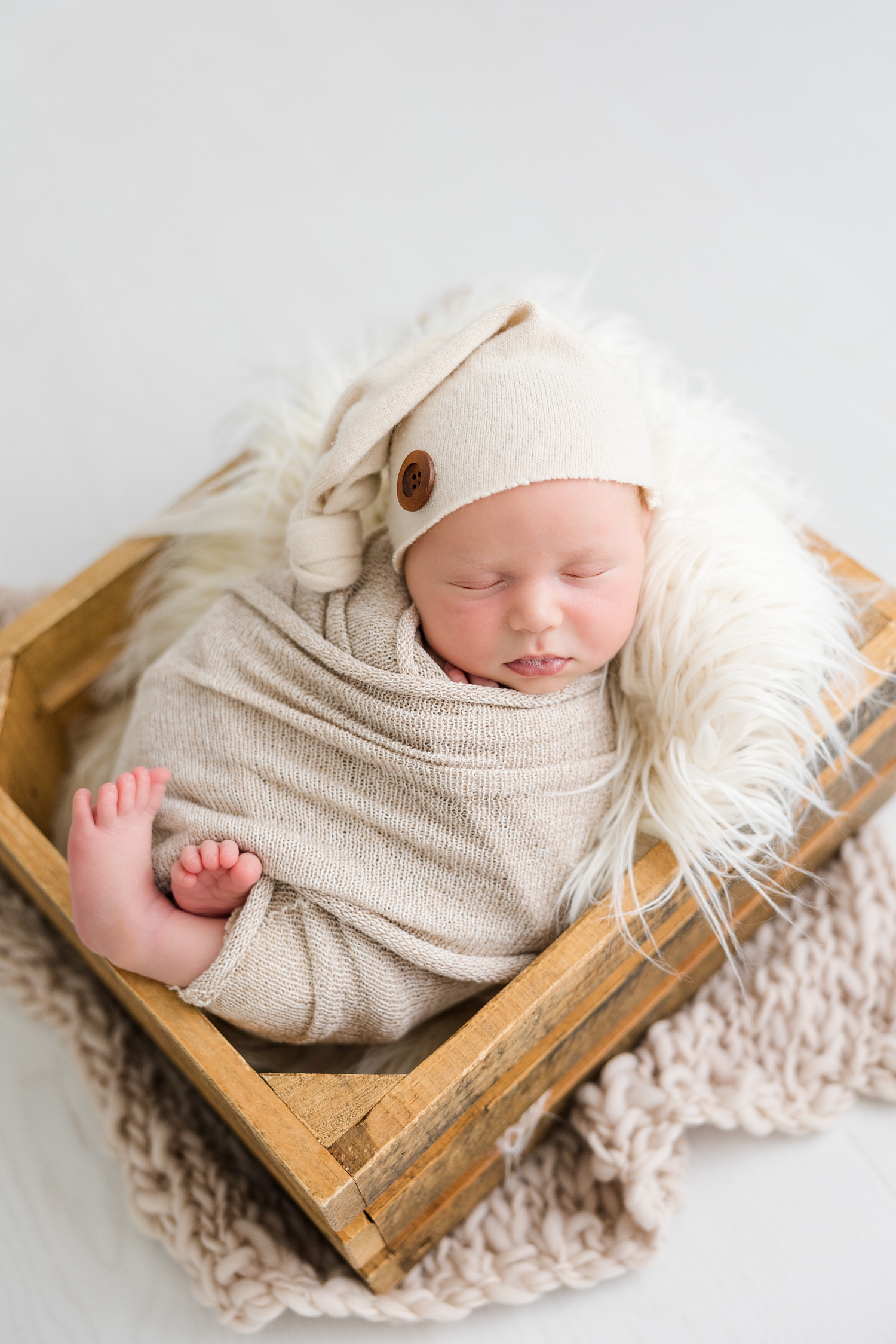 Baby Reed, wrapped in a neutral cloth wearing a neutral night cap, is nestled in a wooden box placed on a white wood floor | CB Studio