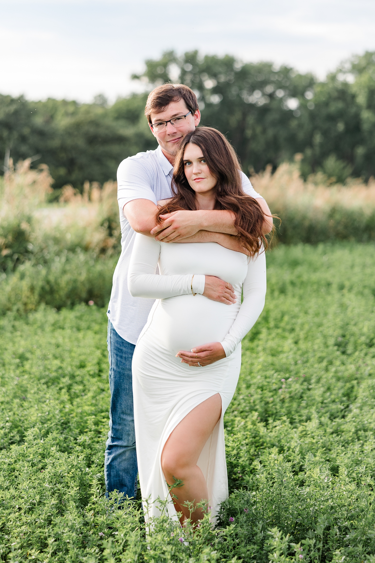 Brady embraces Jenna from behind and Jenna hugs her pregnant belly as they stand in the middle of an alfalfa field | CB Studio