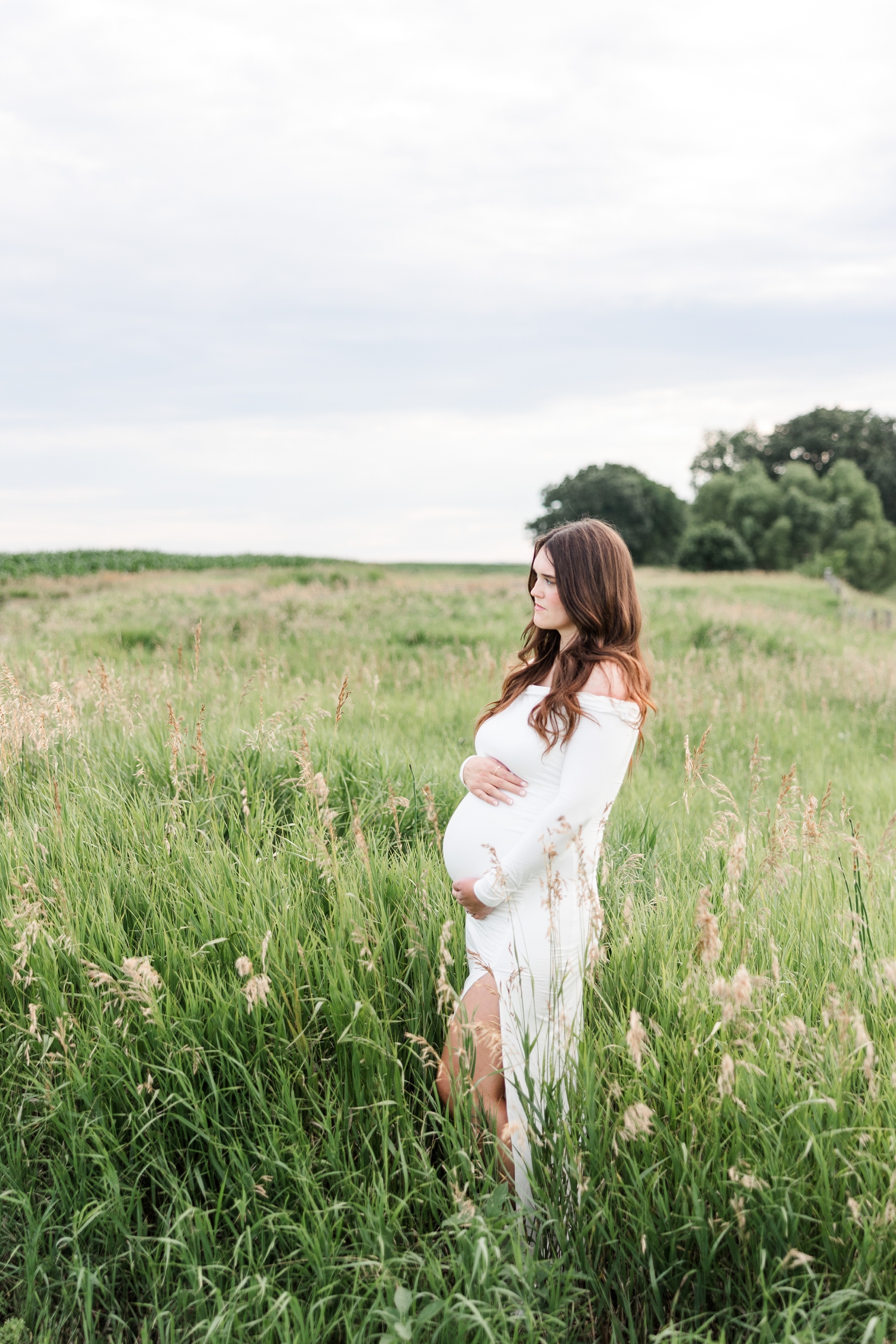 Jenna stands in a grassy pasture as she hugs her pregnant belly and looks off in the distance to the left | CB Studio