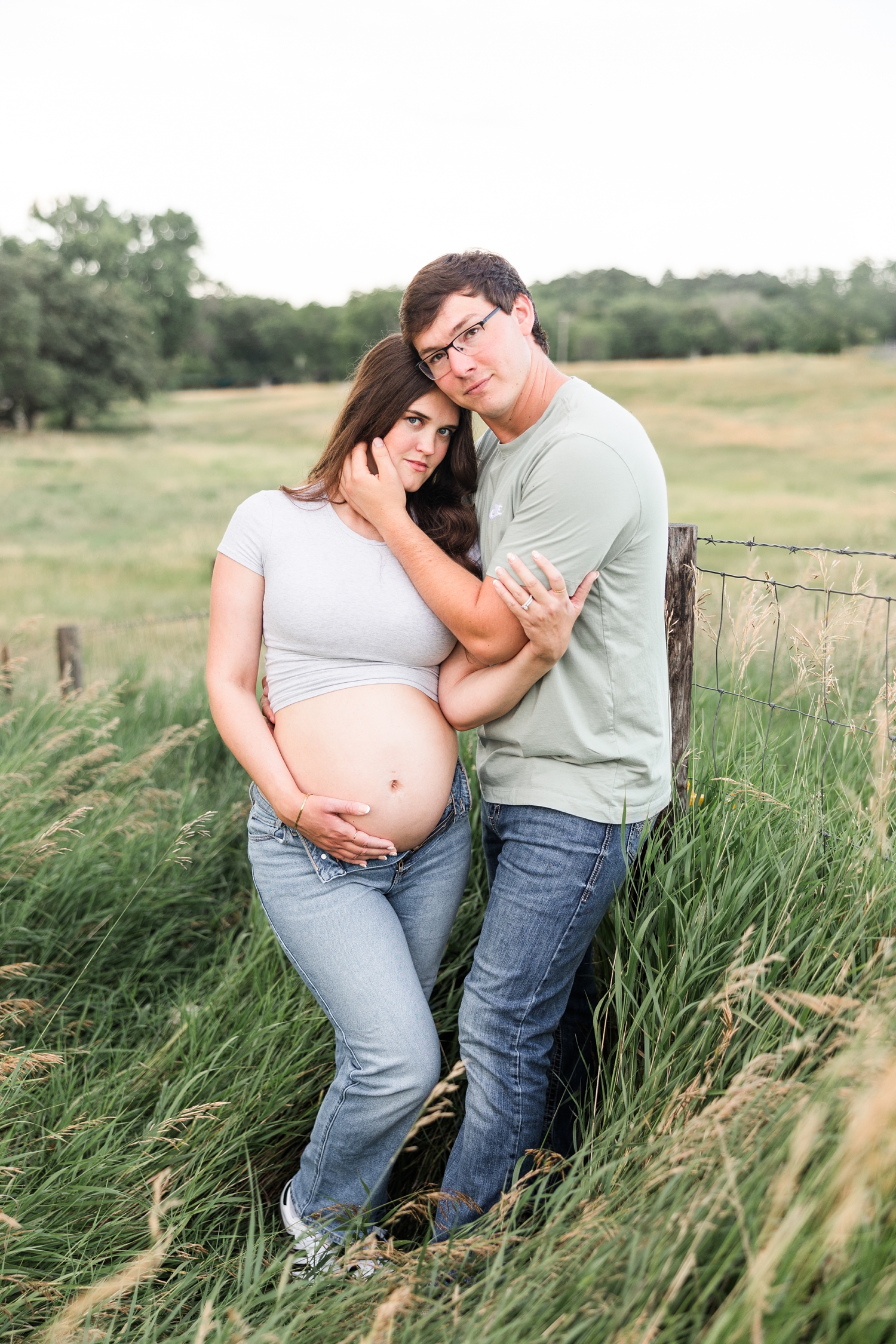 Brady embraces Jenna and her pregnant belly as they stand in the middle of a grassy pasture along a fence line | CB Studio