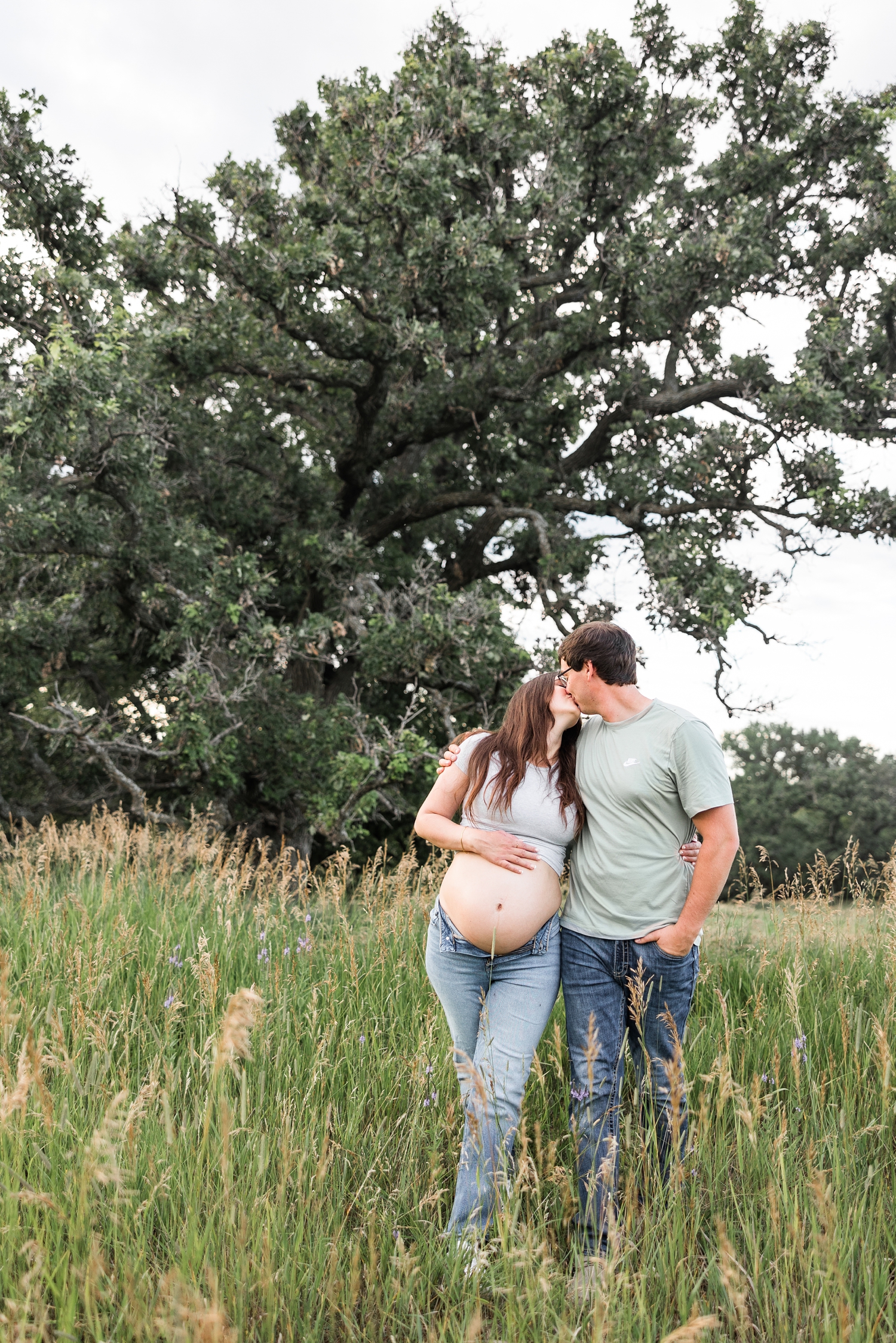 Brady kisses Jenna as they stand in the middle of a grassy pasture with a giant, majestic oak tree in the background and Jenna holds her baby bump | CB Studio