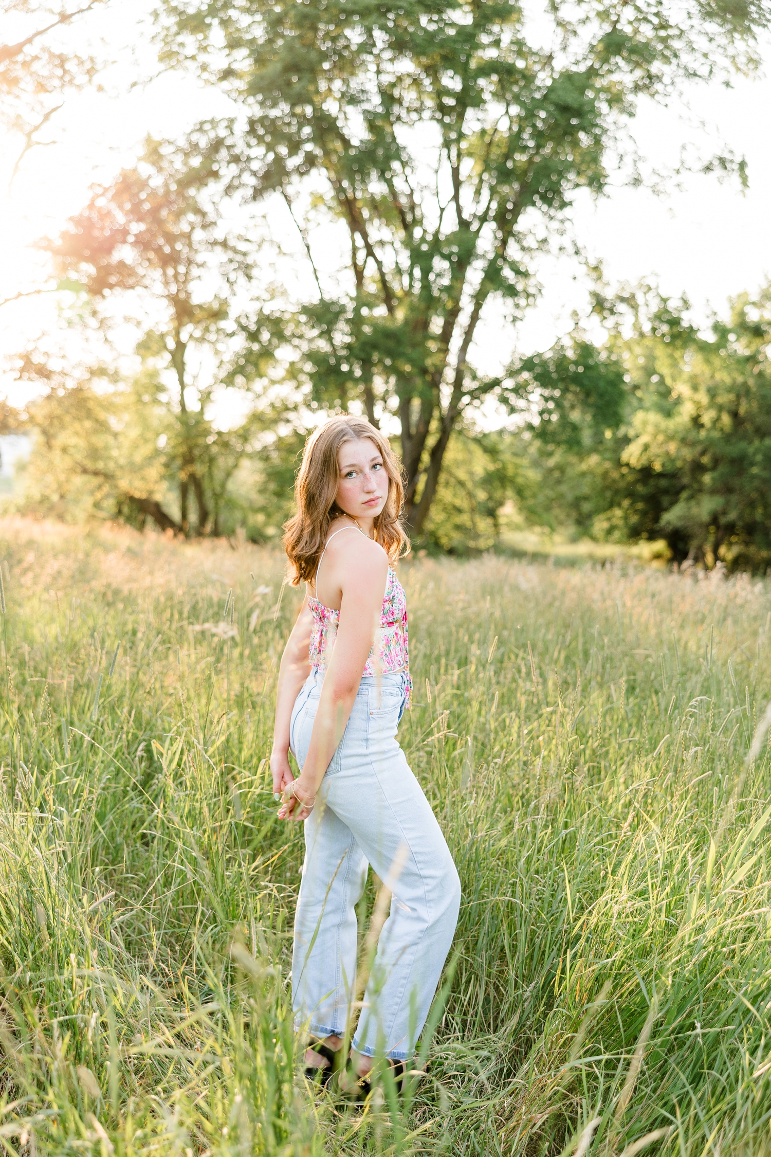 Bre, wearing a floral tank and jeans, stands in a grassy pasture with her hands behind her back | CB Studio