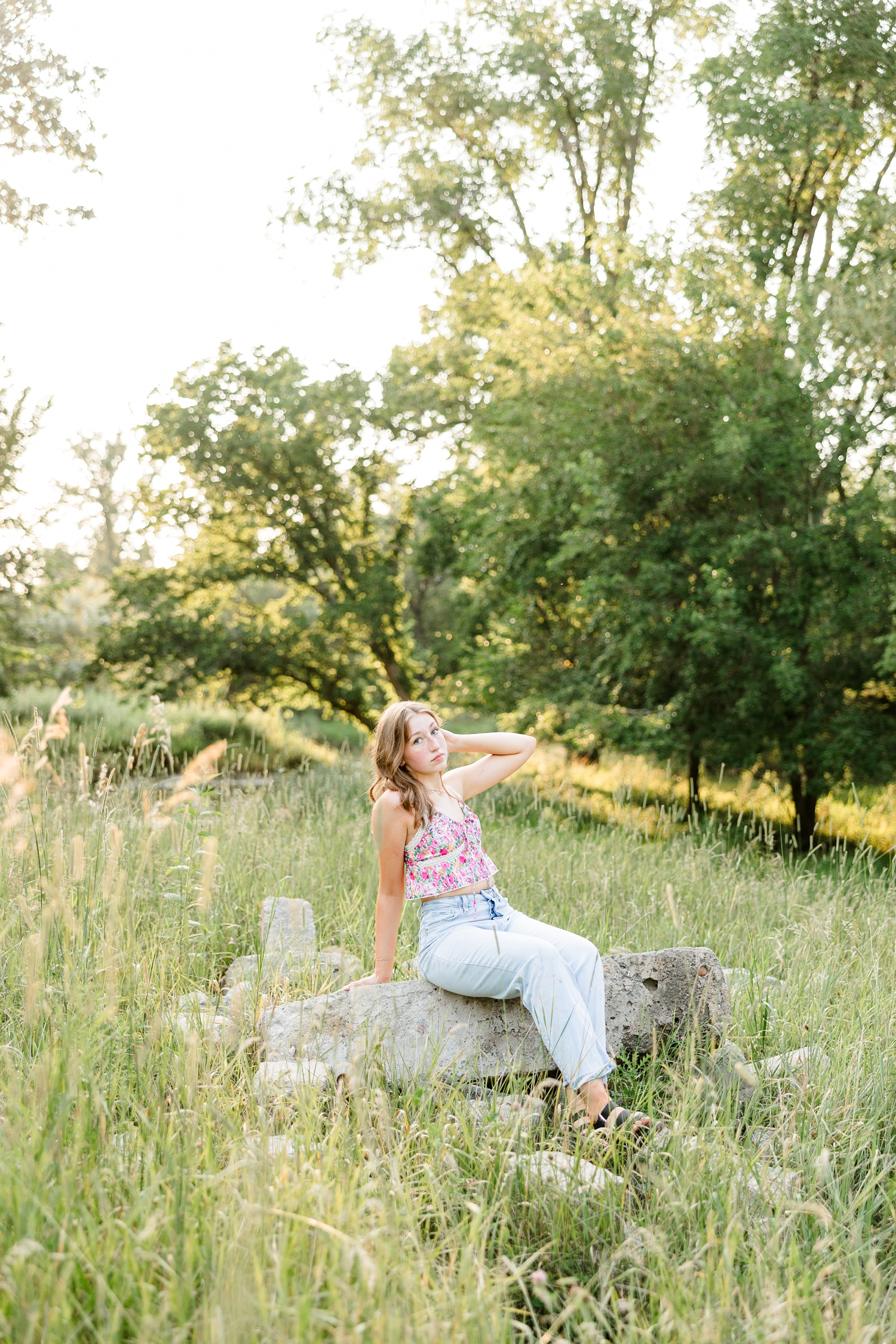 Bre, wearing a floral tank and jeans, sits on some stones in a grassy pasture as she fluffs her hair | CB Studio