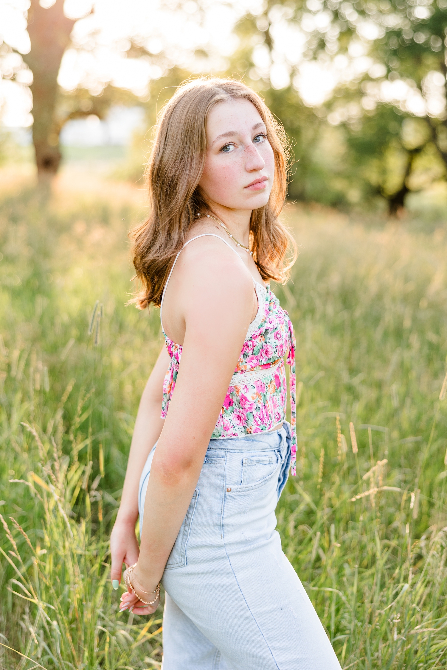 Bre, wearing a floral tank and jeans, stands in a grassy pasture with her hands behind her back | CB Studio