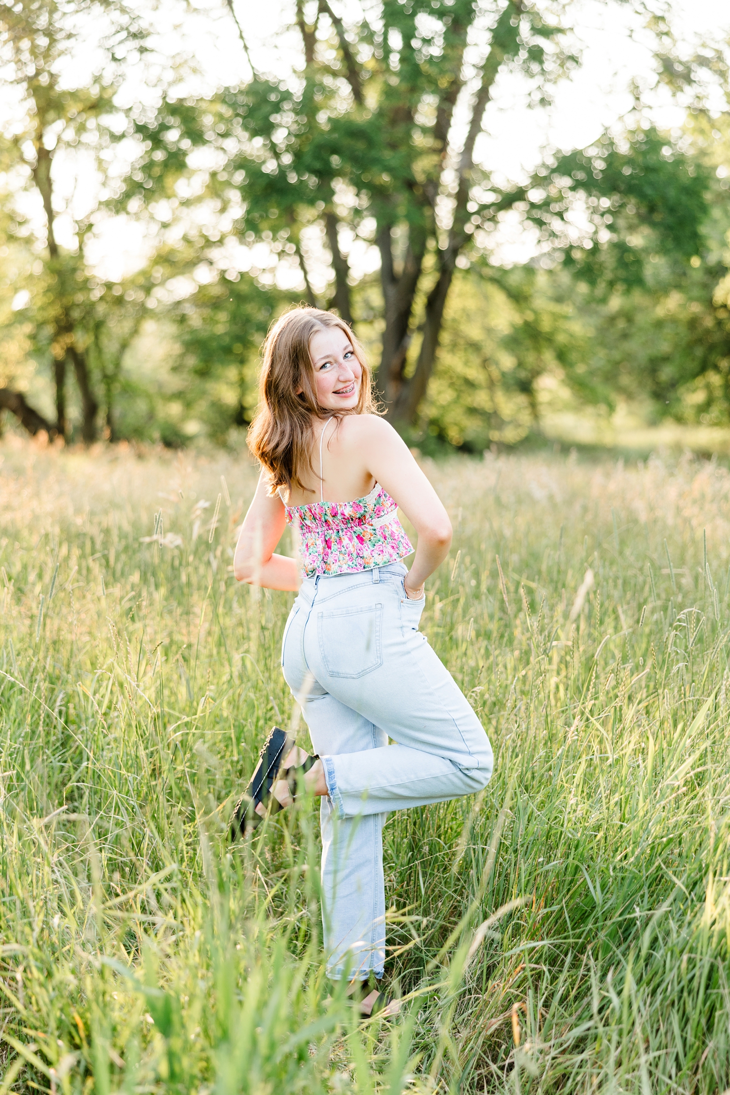 Bre, wearing a floral tank and jeans, stands in a grassy pasture and kicks up her leg | CB Studio