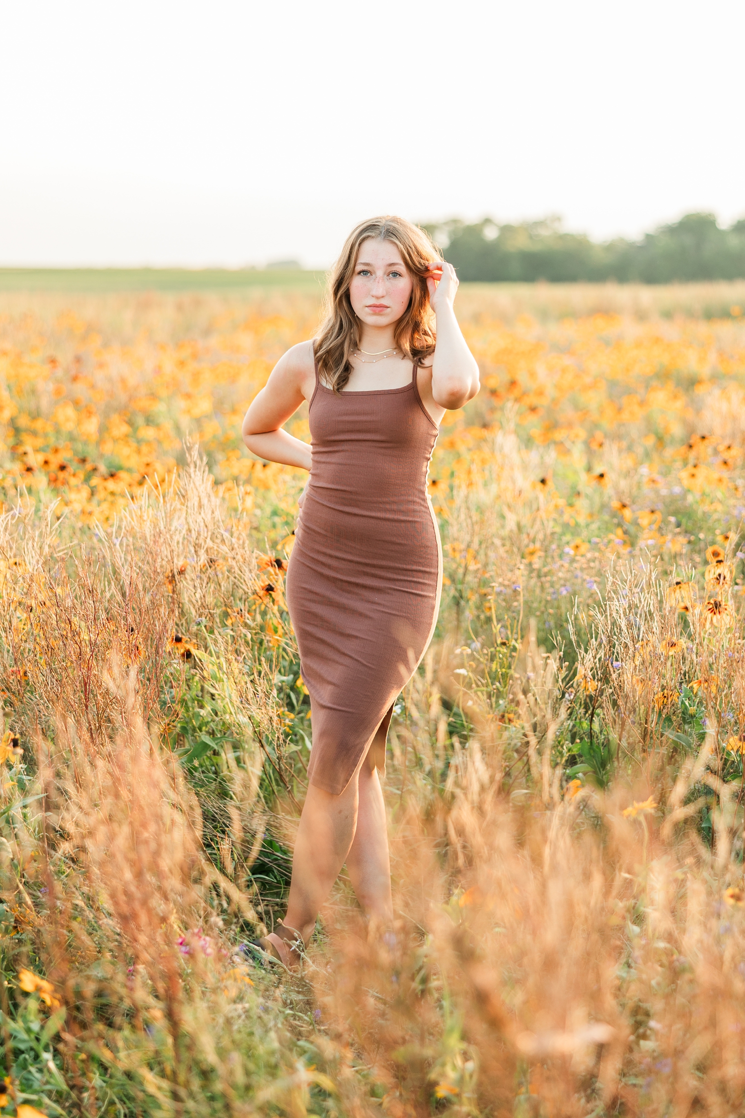 Bre, wearing a brown body con dress, stands in a wildflower field as she tucks her hair behind her ear at golden hour | CB Studio