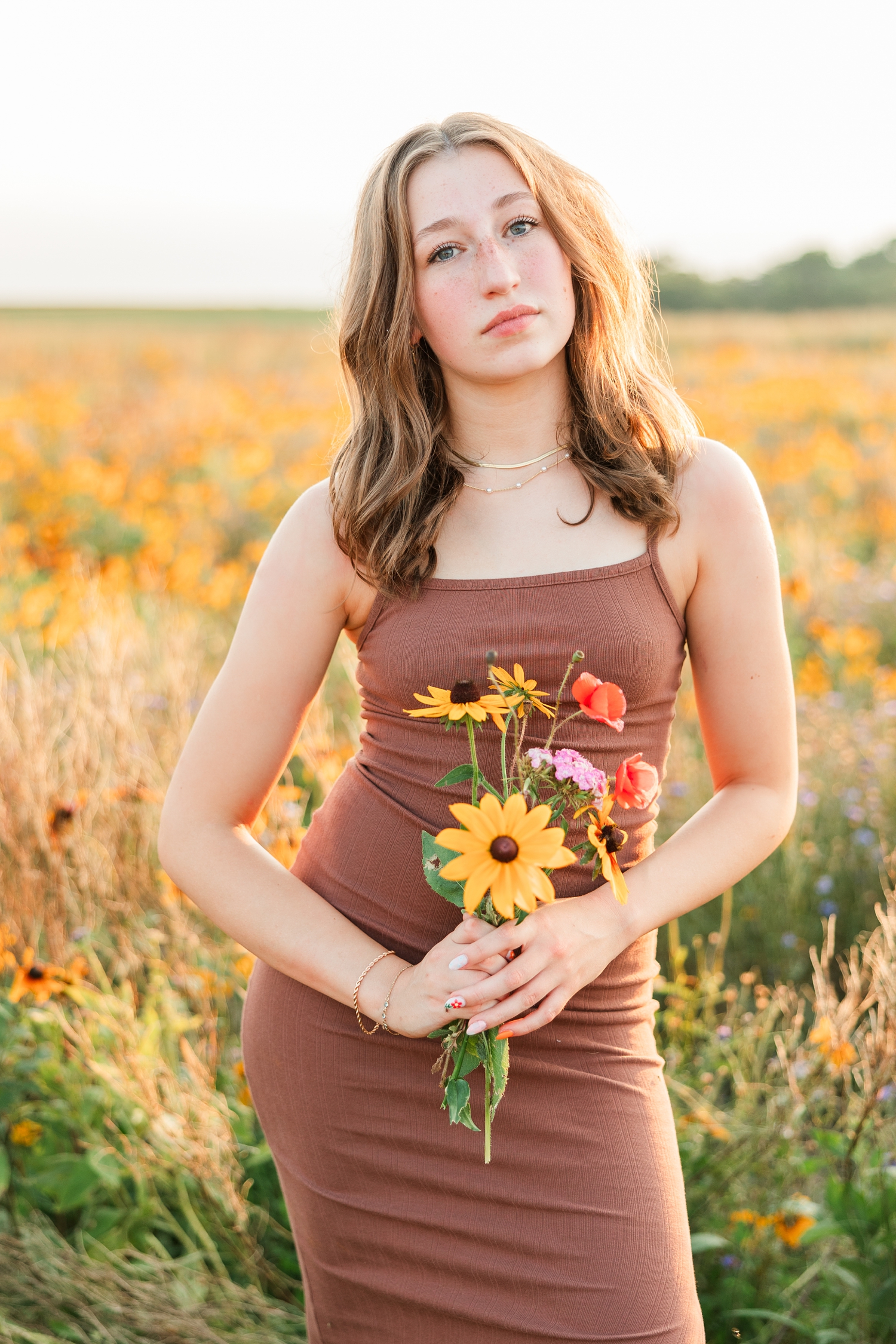 Bre, wearing a brown body con dress, stands in a wildflower field holding a small bouquet at golden hour | CB Studio