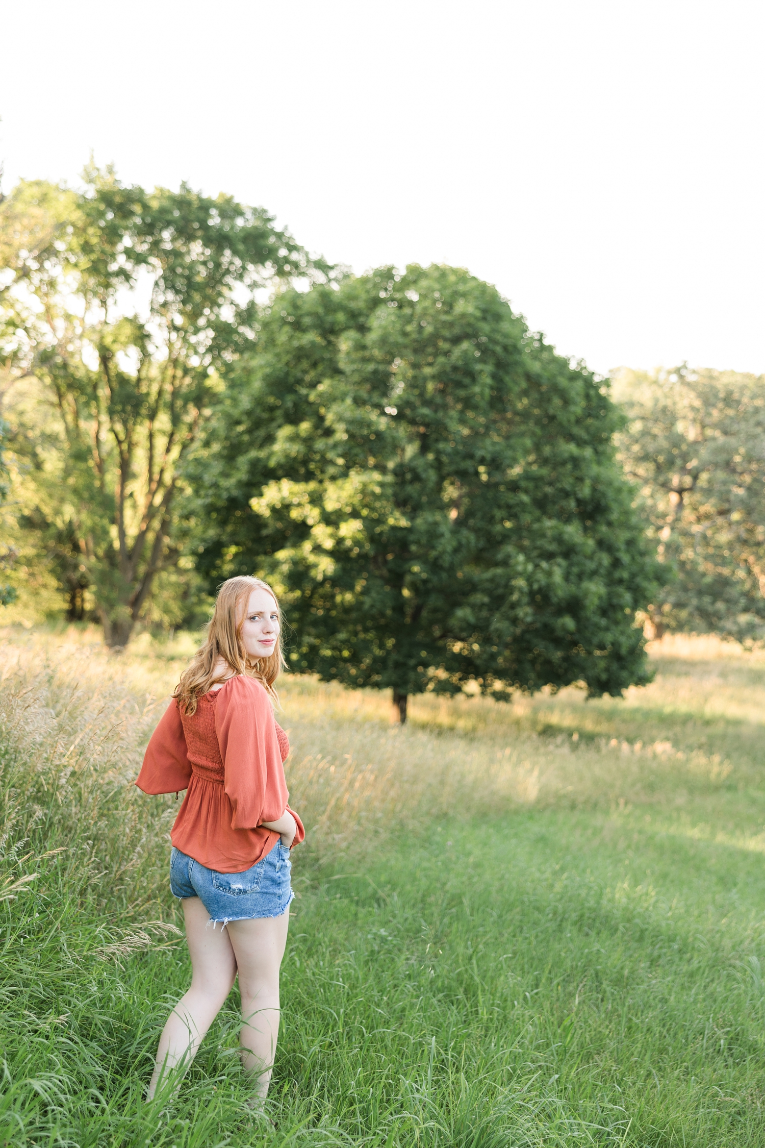 Hannah, wearing denim shorts with a rust colored peasant top, walks with her hands in her pockets in a grassy pasture while looking back over her shoulder | CB Studio