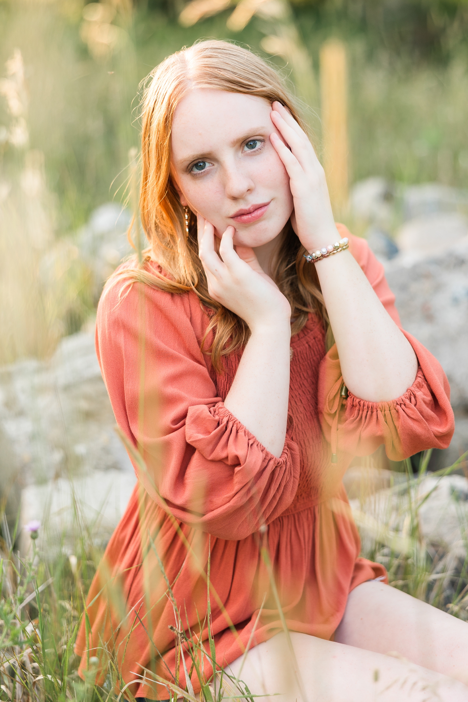 Hannah, wearing denim shorts with a rust colored peasant top, sits on a stone surrounded by tall grasses in a grassy pasture while cupping her face with her hands | CB Studio