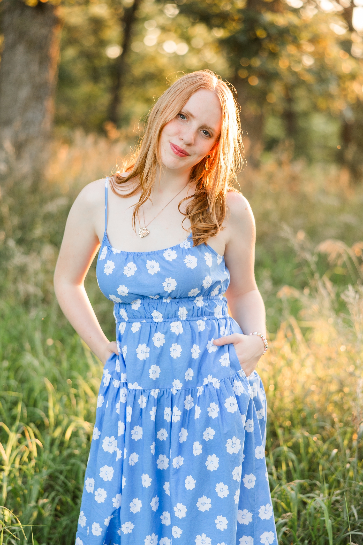 Hannah, wearing a blue dress with white flowers, stands with her hands in her pockets in a grassy pasture surrounded by oak trees | CB Studio