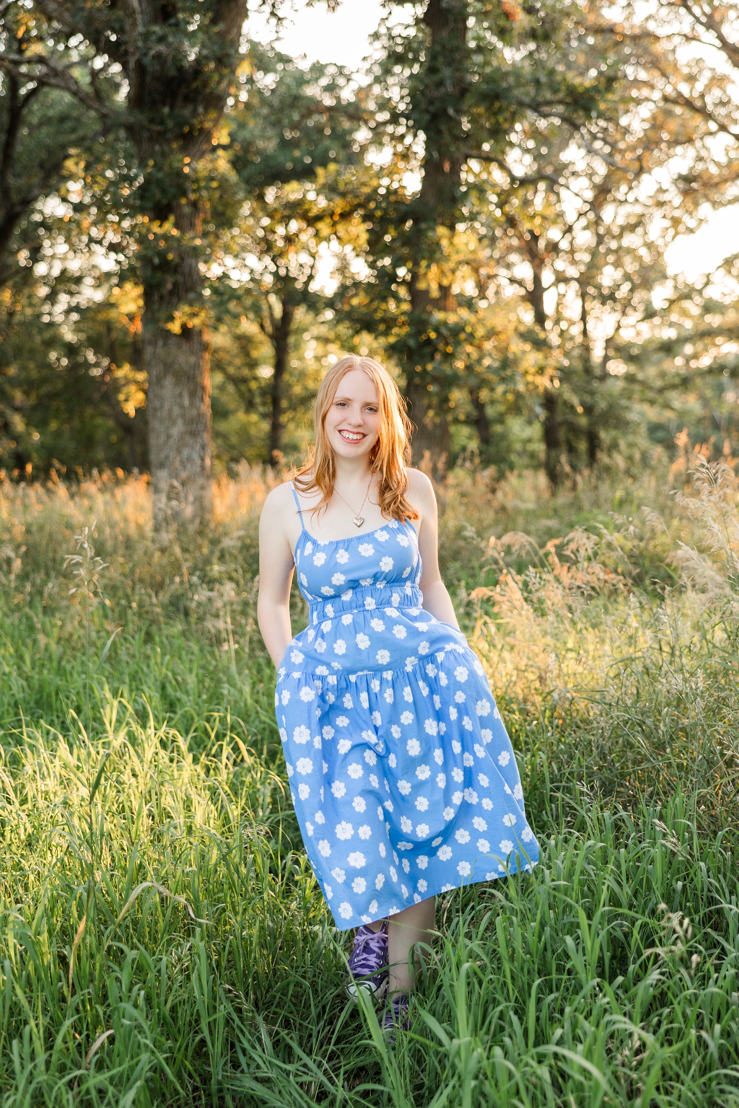 Hannah, wearing a blue dress with white flowers, walks with her hands in her pockets in a grassy pasture surrounded by oak trees | CB Studio