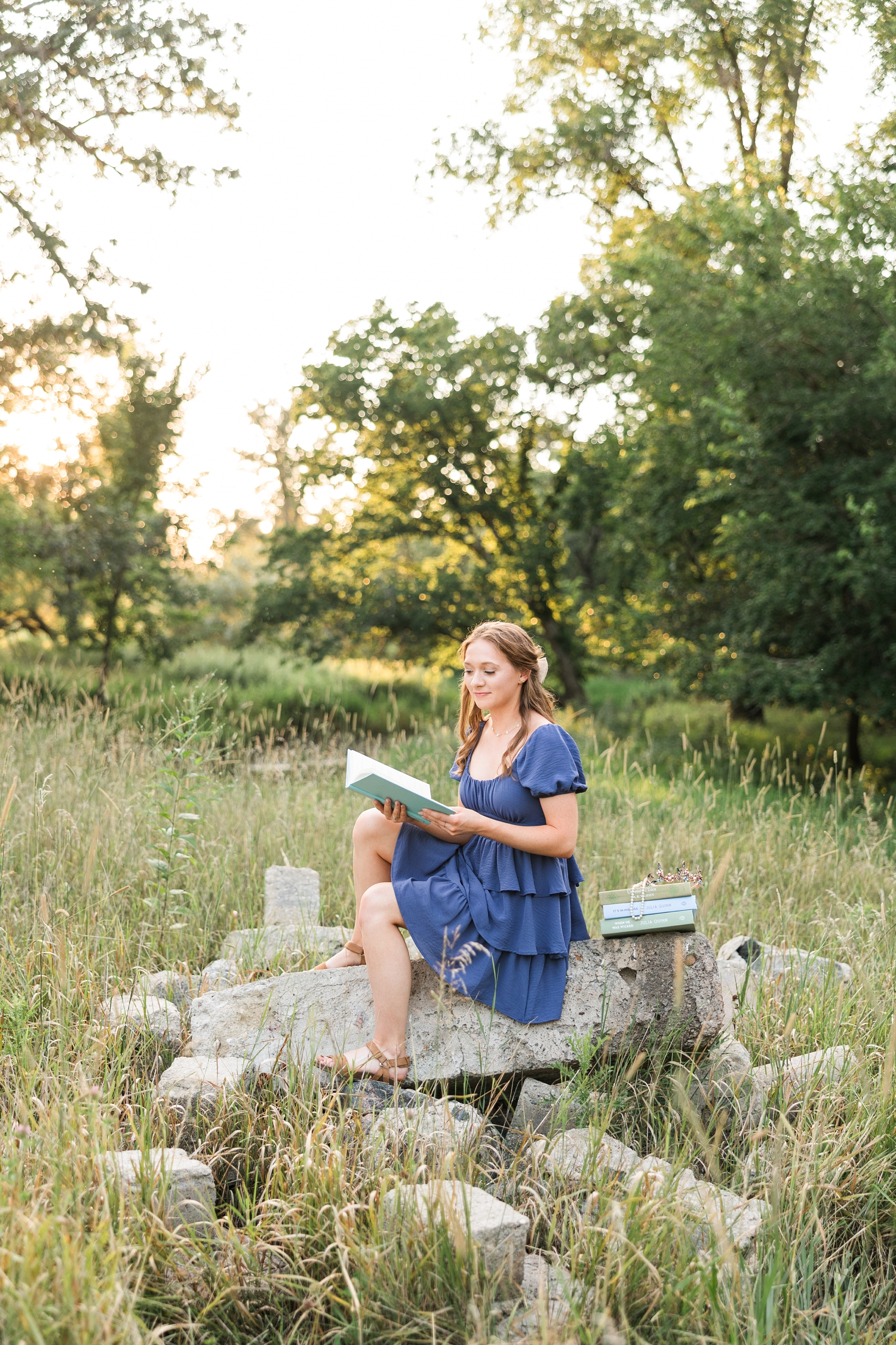 Izzie sits on a pile of stones in a grassy pasture as she reads her Bridgerton books | CB Studio