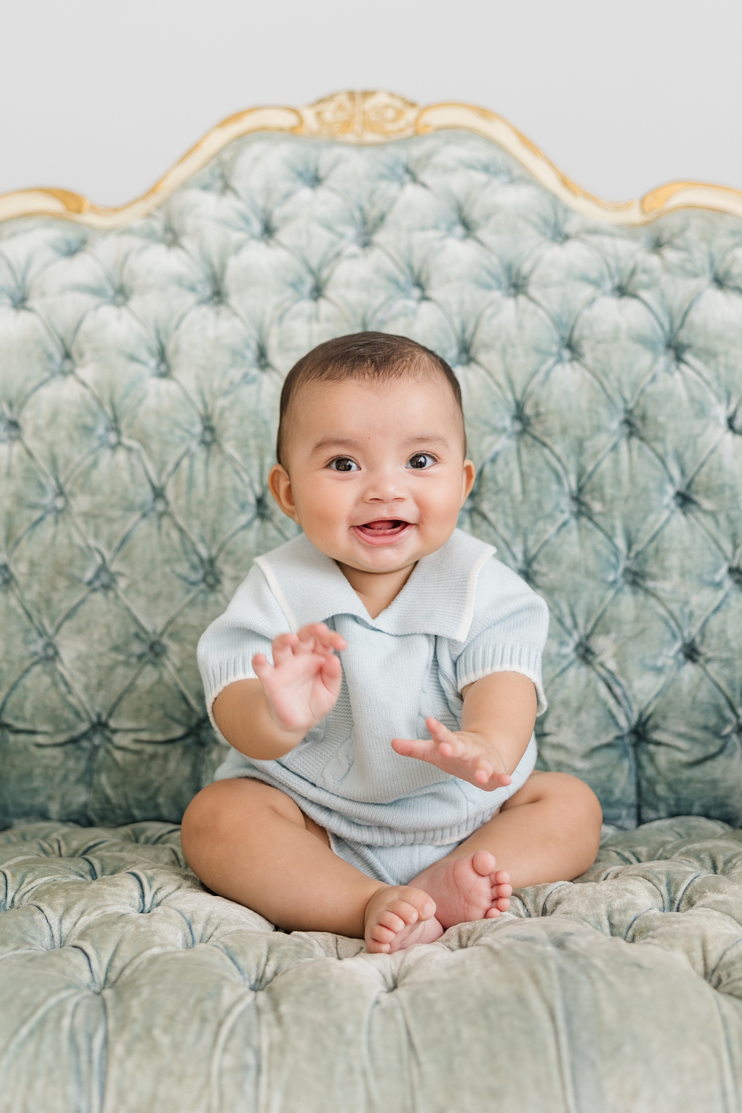 Baby Zachary, dressed in a light blue knit, vintage matching set, smiles as he waves his hands while sitting on a light blue victorian tufted couch with gold inlay detail | CB Studio