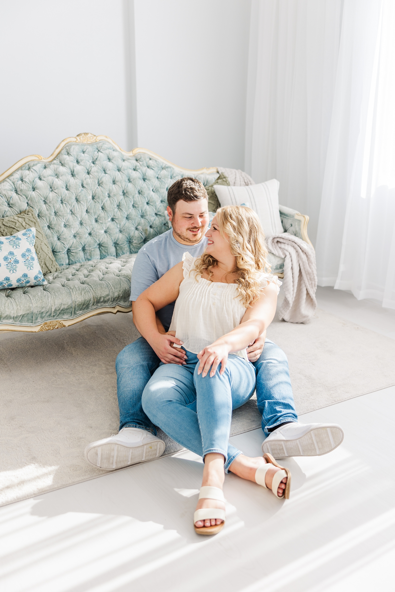 Connor sits on the floor of an all white room leaning against a vintage victorian tufted couch while Natalie sits in between is legs and smile at each other | CB Studio