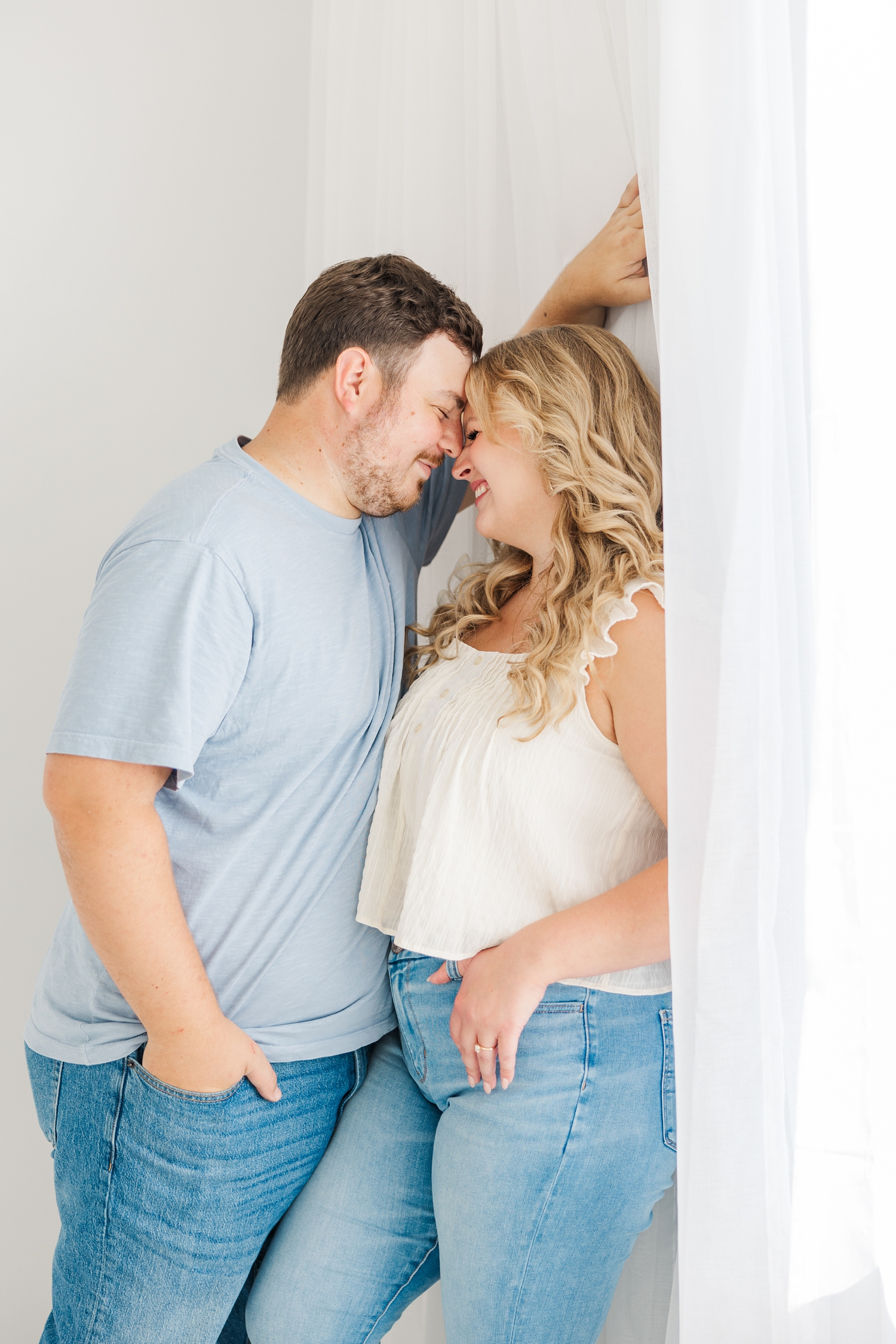 Natalie leans against a white wall with white curtains as Connor leans towards her and they both touch noses | CB Studio