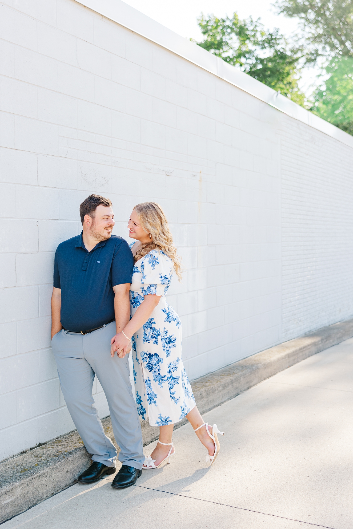 Connor leans his shoulder on the side of a light blue brick building in downtown Algona, IA while Natalie holds his hand and they smile at each other | CB Studio