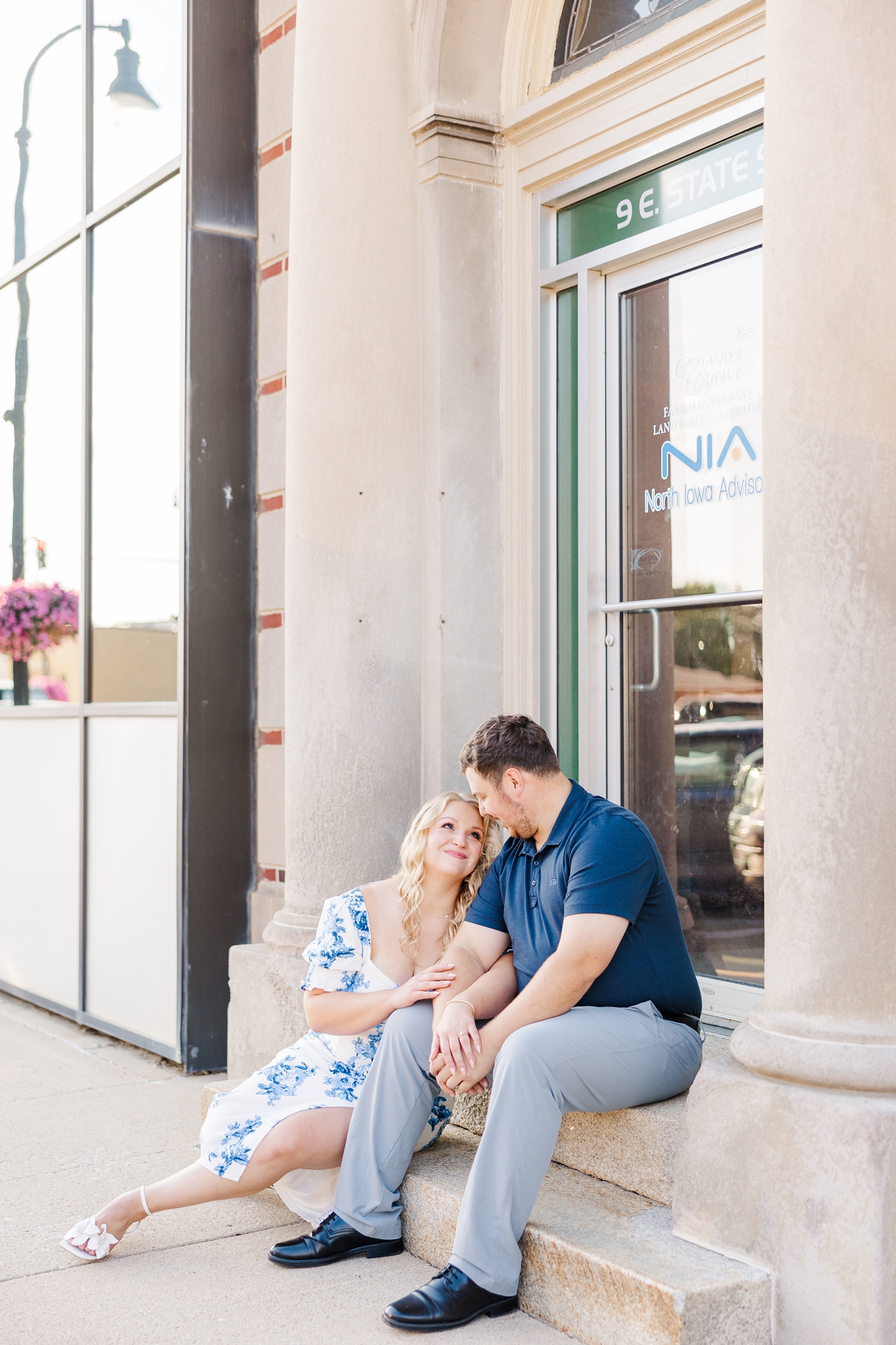 Connor and Natalie smile at each other while sitting on the steps of a historic stone building in downtown Algona, IA | CB Studio