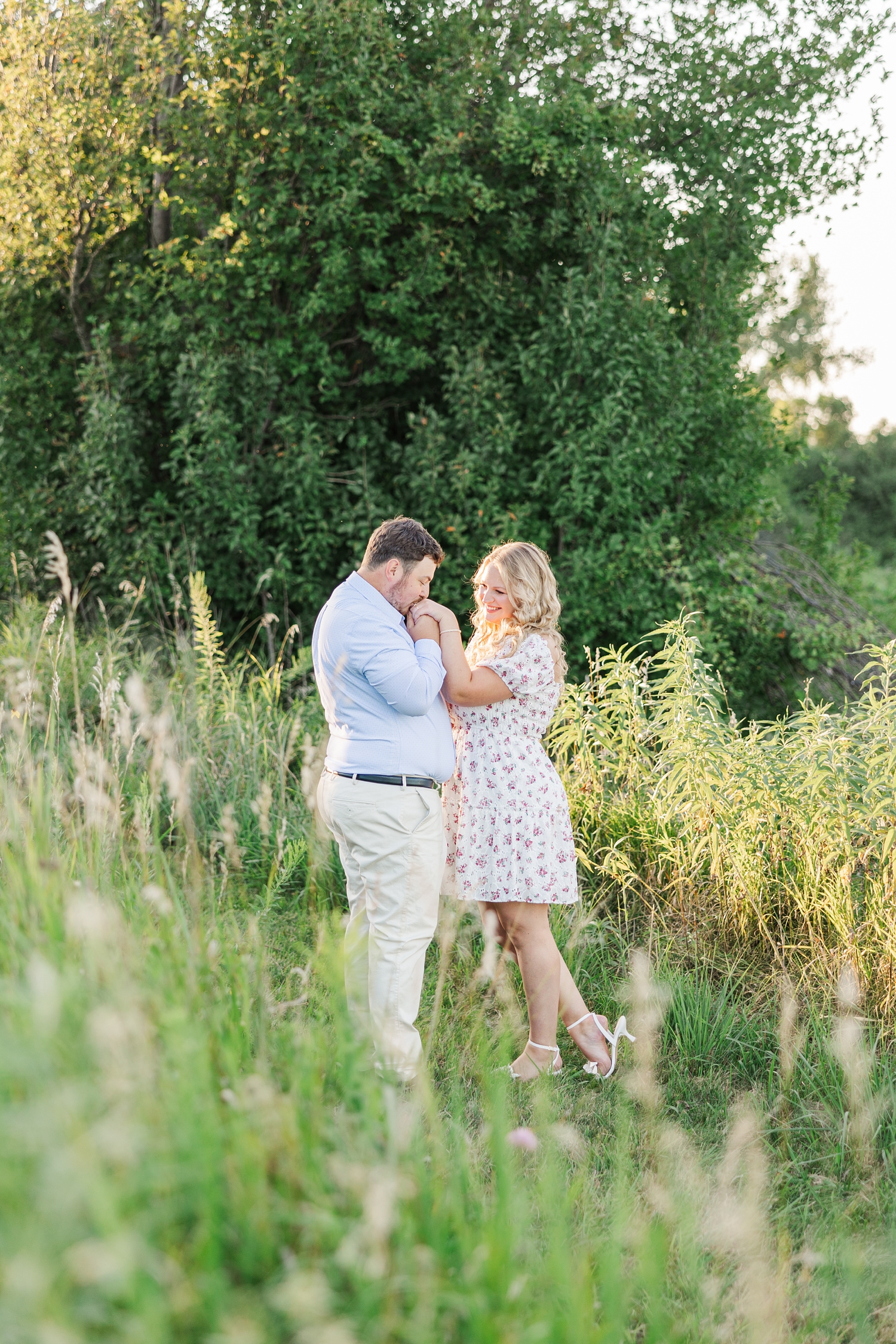 Connor pulls Natalie's hand up and kisses it while standing in a grassy pasture at Water's Edge Nature Center | CB Studio