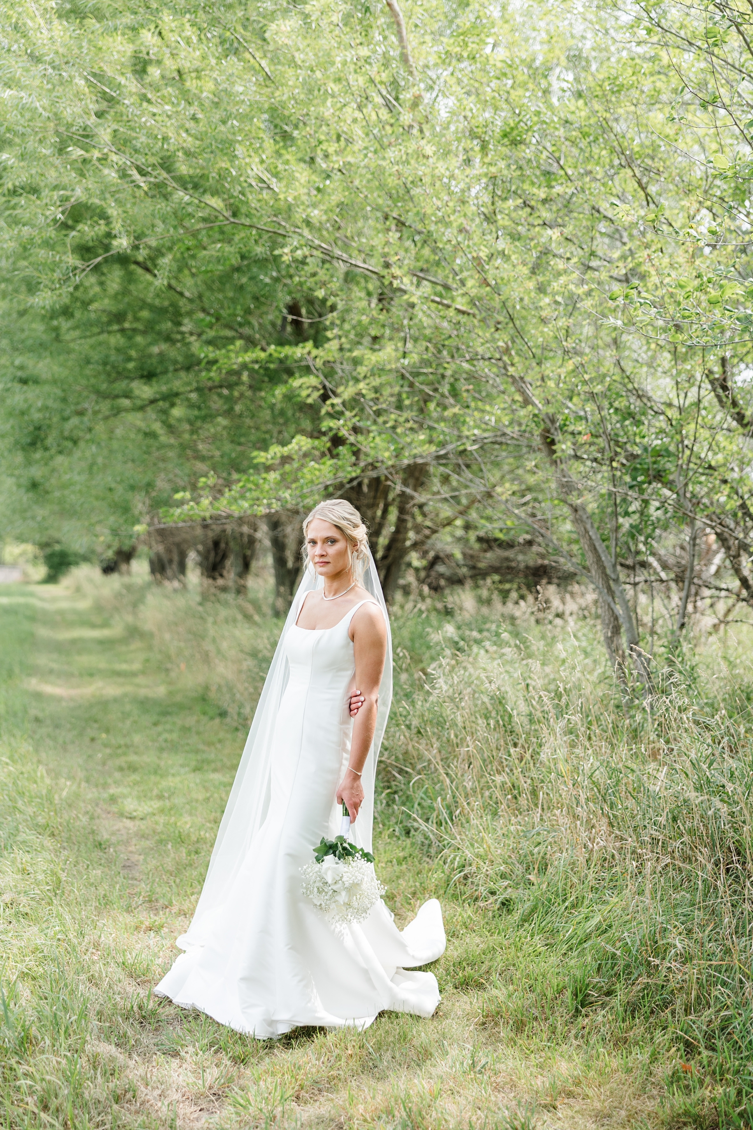 Bride, Nicole, stands in a walking path in a grassy pasture on her farm in northern Iowa holding her white rose and baby's breath bouquet | CB Studio