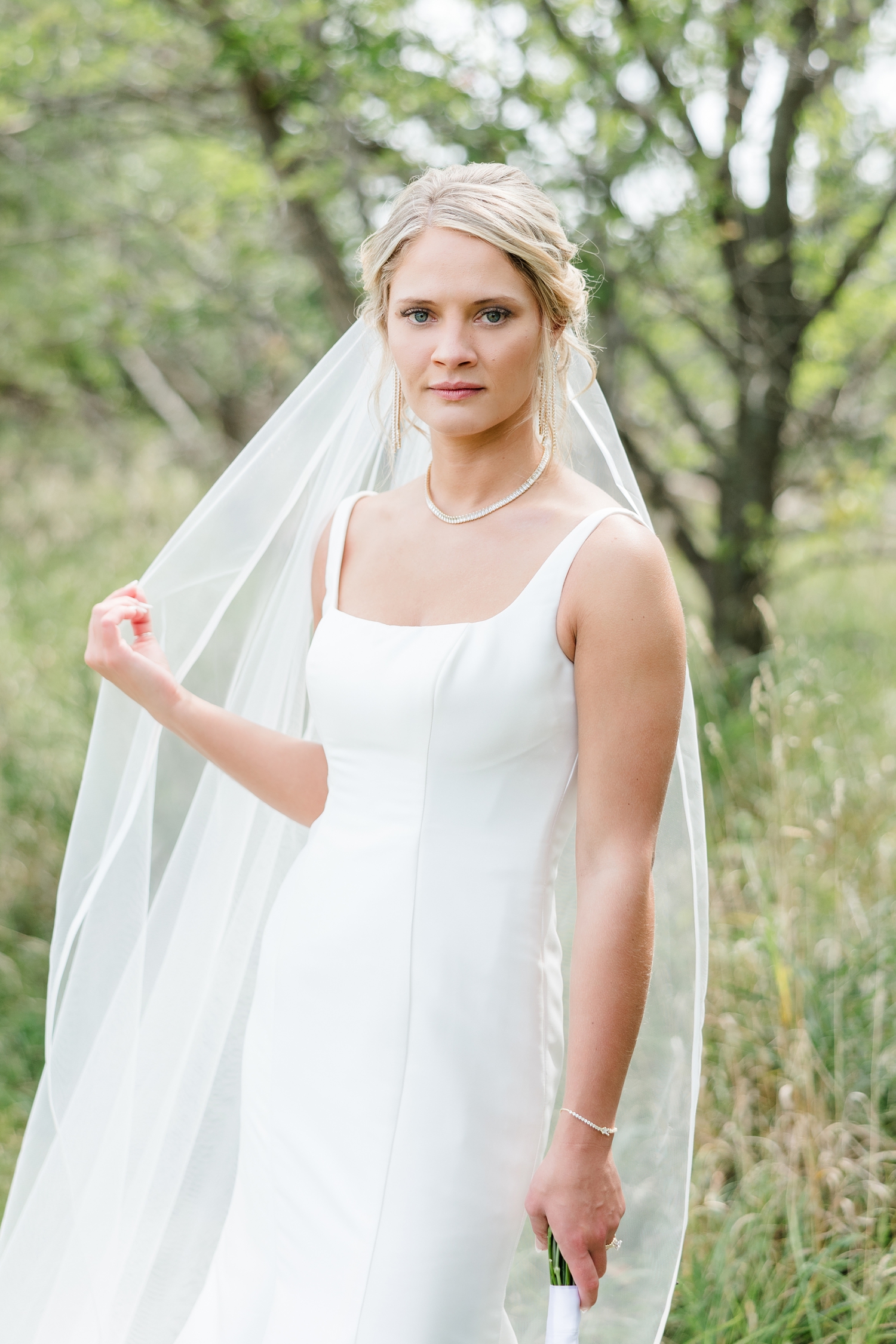 Bride, Nicole, stands in a grassy pasture on her farm in northern Iowa as she slowly runs her finger down her veil | CB Studio