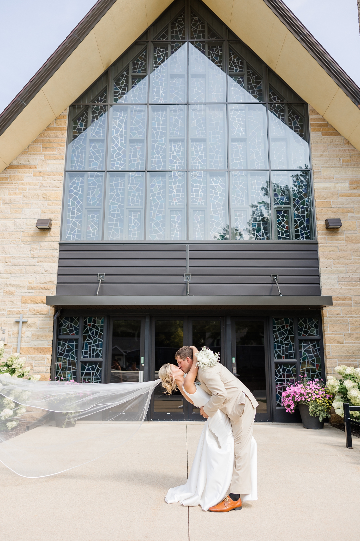 Reed dips his new bride for a kiss while her veil flys in the air to the left as they stand in front of St. Mary's Catholic church in Humboldt, IA | CB Studio