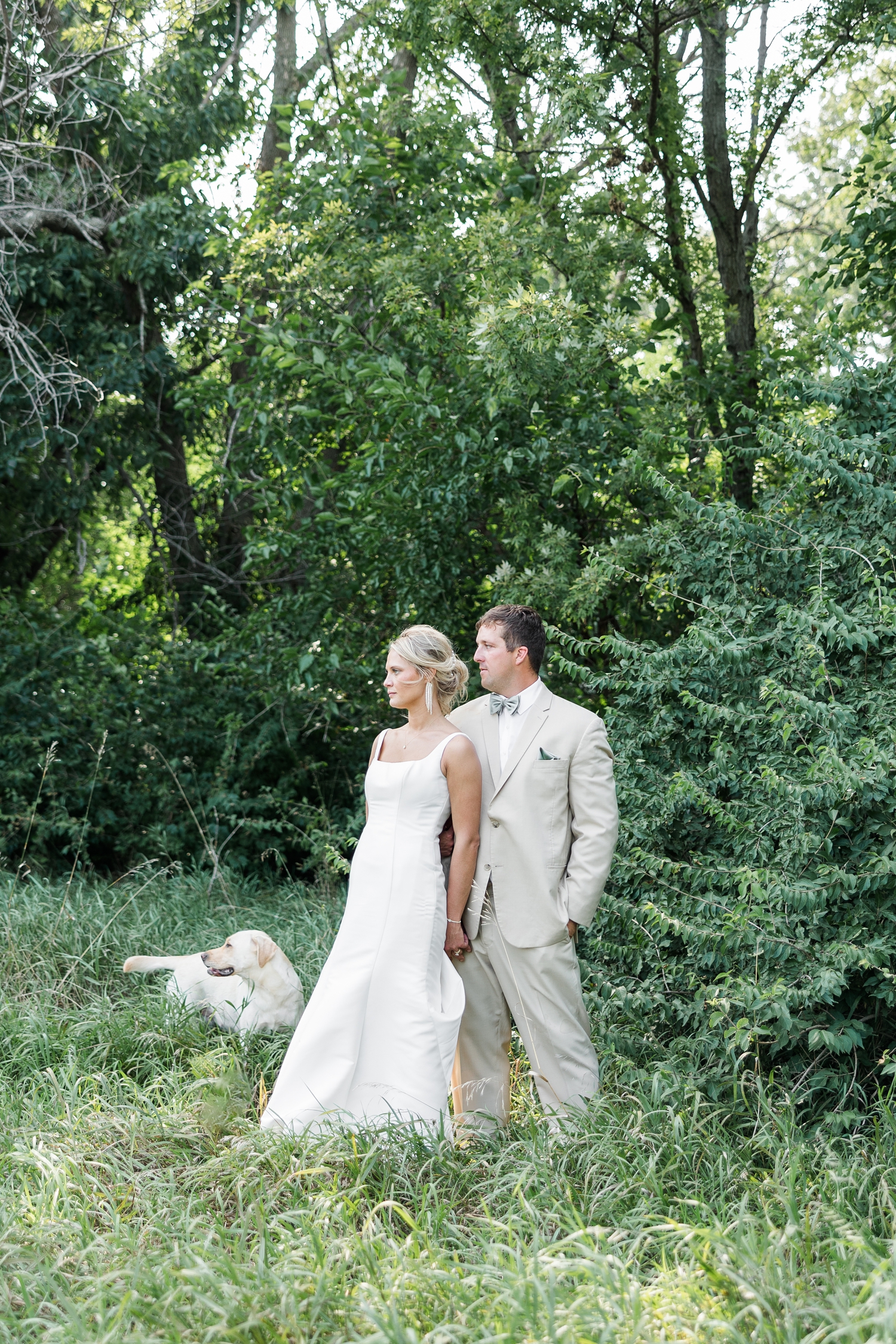 Nicole leans on her groom's shoulder as they both look to the left while standing in a grassy field in a pasture on their farm. Their golden retriever, Nala, stands slightly in the background to the left while also looking left. | CB Studio