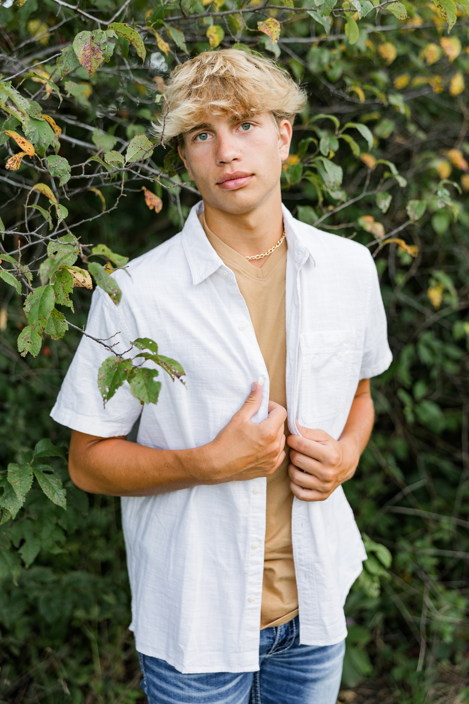 Tyler holds the open panels of his button down shirt while standing in front of a bush starting to turn yellow for fall | CB Studio