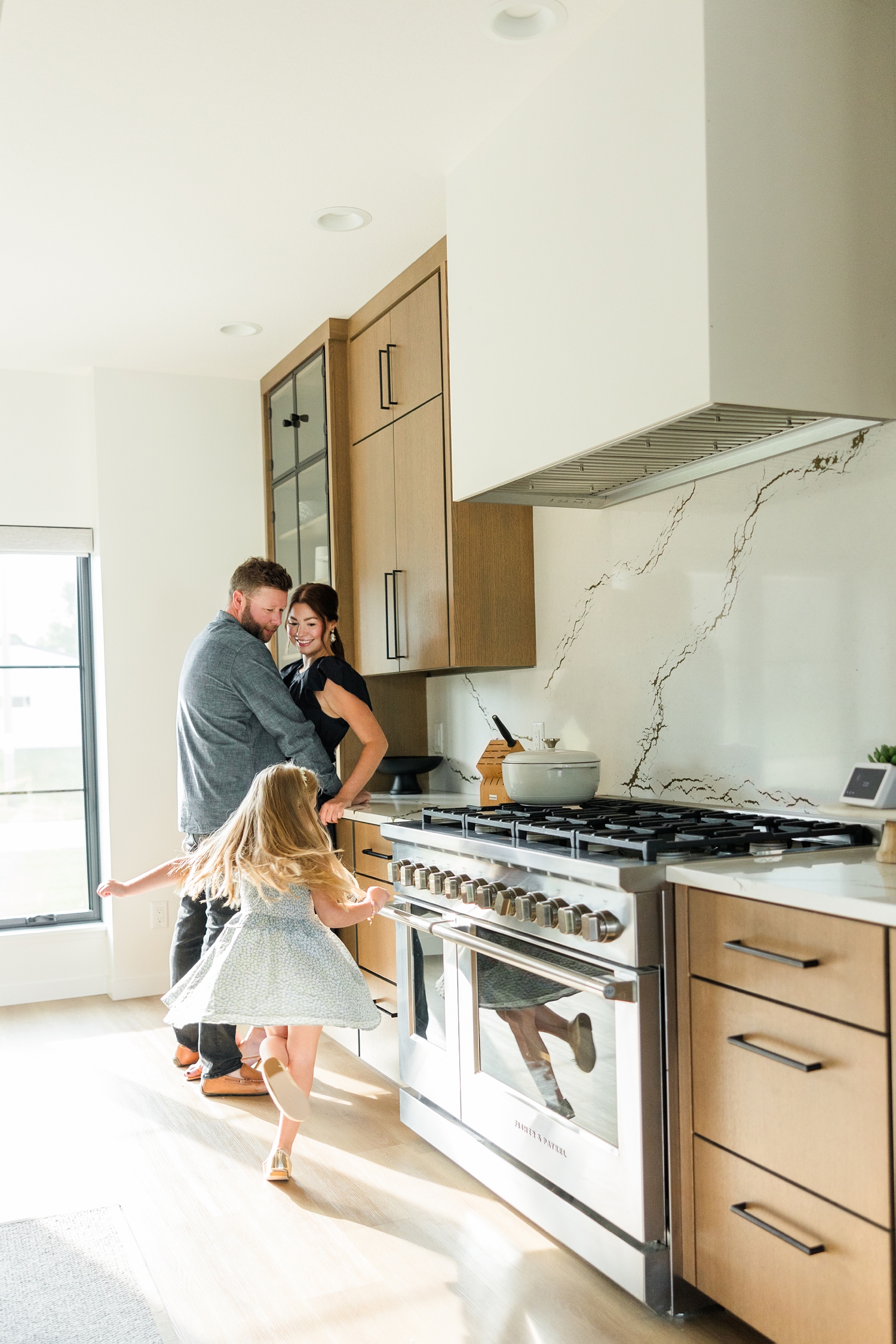 The Arndorfers stand in their kitchen while their daughter twirls around in the sunlight in their new, modern home for an in-home lifestyle family photoshoot | CB Studio