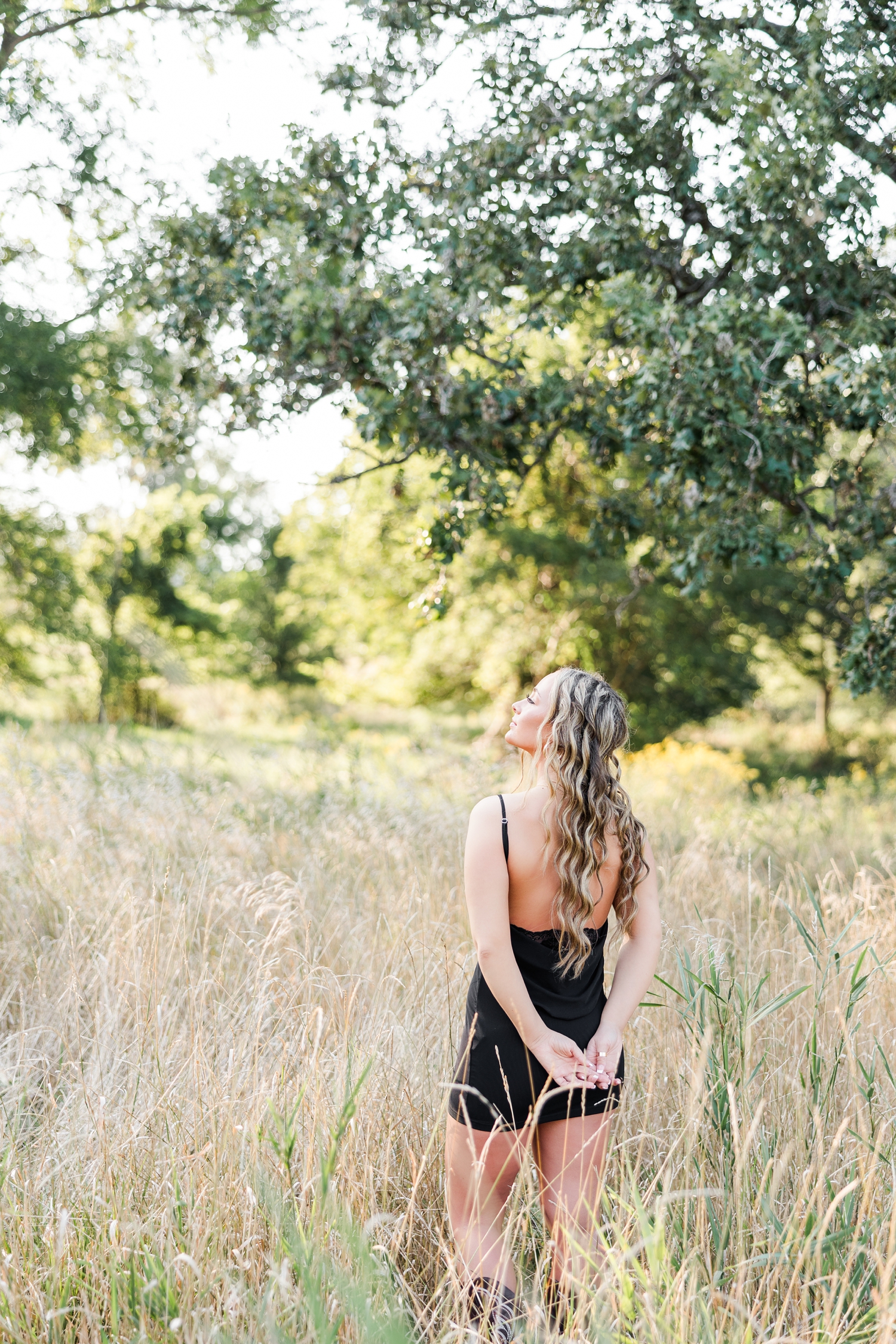 Blaire, wearing a black mini dress, stands in the middle of a grassy field with her back towards the camera as she looks up to the sky | CB Studio