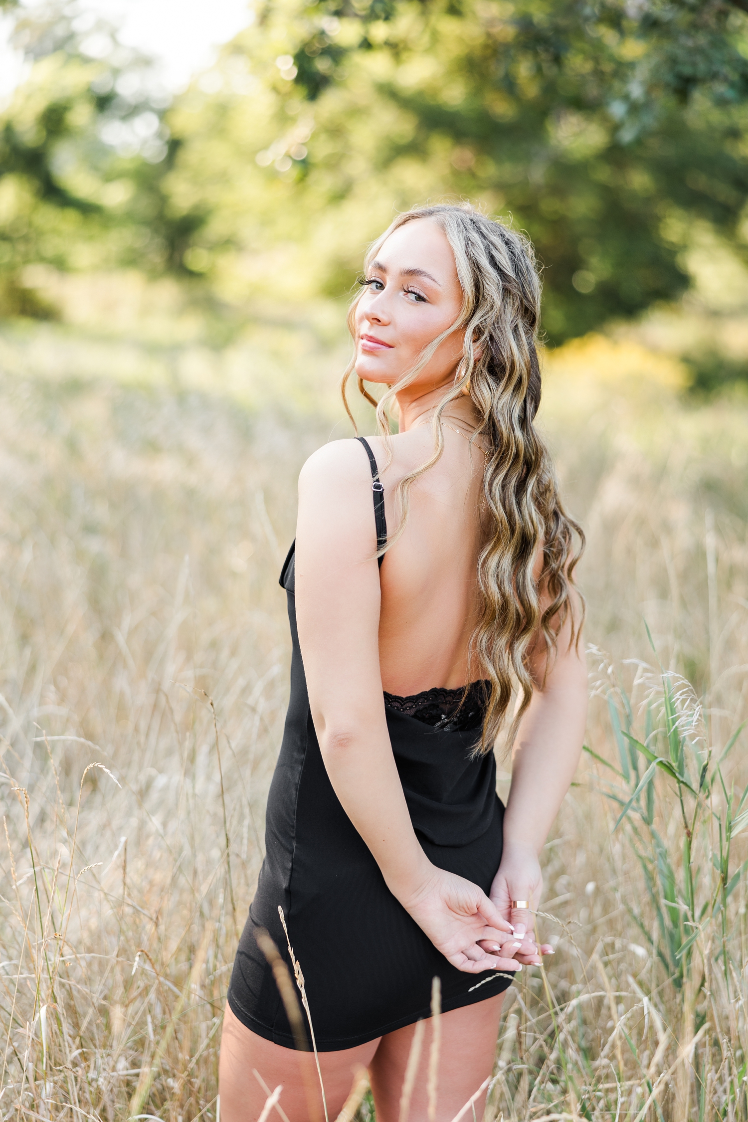 Blaire, wearing a black mini dress, stands in the middle of a grassy field with her back towards the camera as she looks over her left shoulder | CB Studio