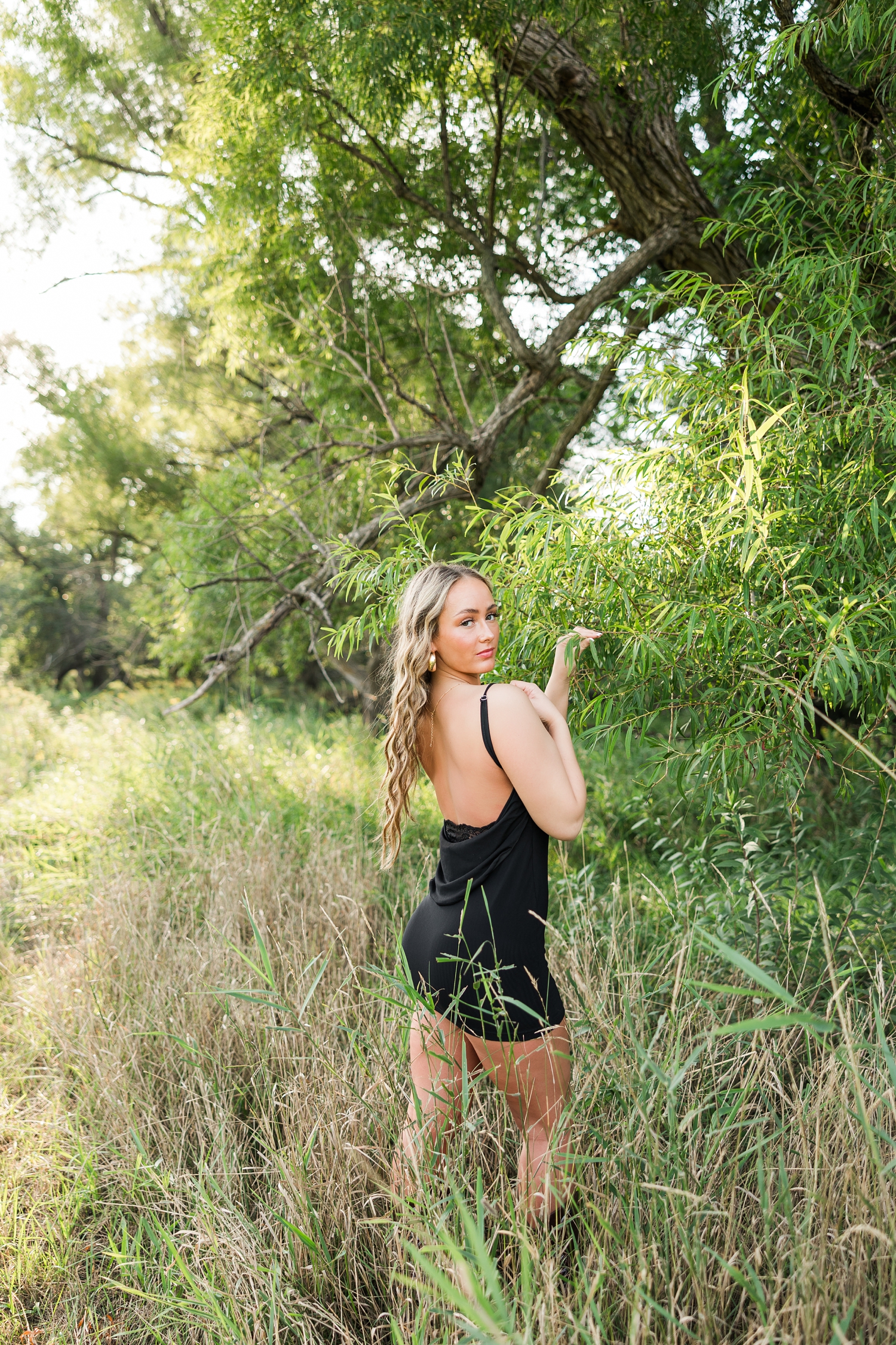 Blaire, wearing a black mini dress, stands in the middle of a grassy field holding the branch of a tree as she looks over her right shoulder | CB Studio