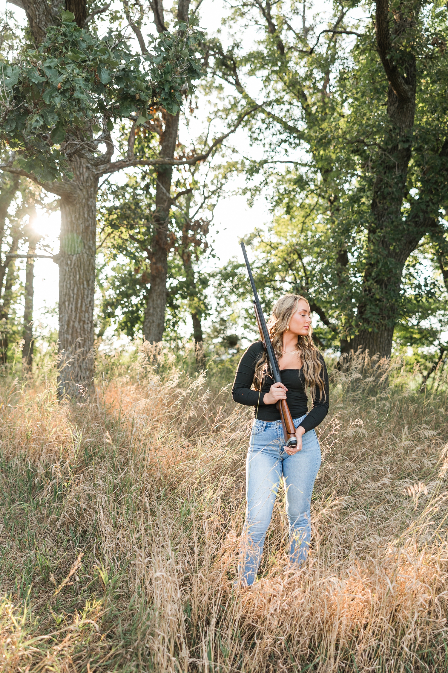 Blaire, wearing jeans and a black long sleeve top, stands in the middle of a grassy field holding her trap shooting rifle over her right shoulder as she stares to the left | CB Studio