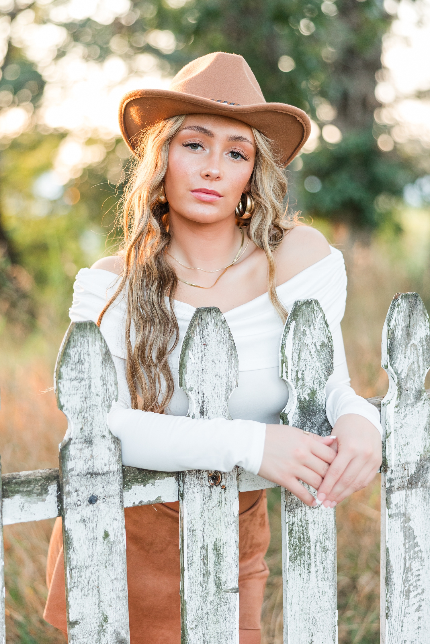 Blaire, wearing a brown suede mini skirt, white off the shoulder long sleeve top and brown suede cowboy hat, stands with her arms gently wrapped around an old rustic white picket fence in a grass field during golden hour | CB Studio