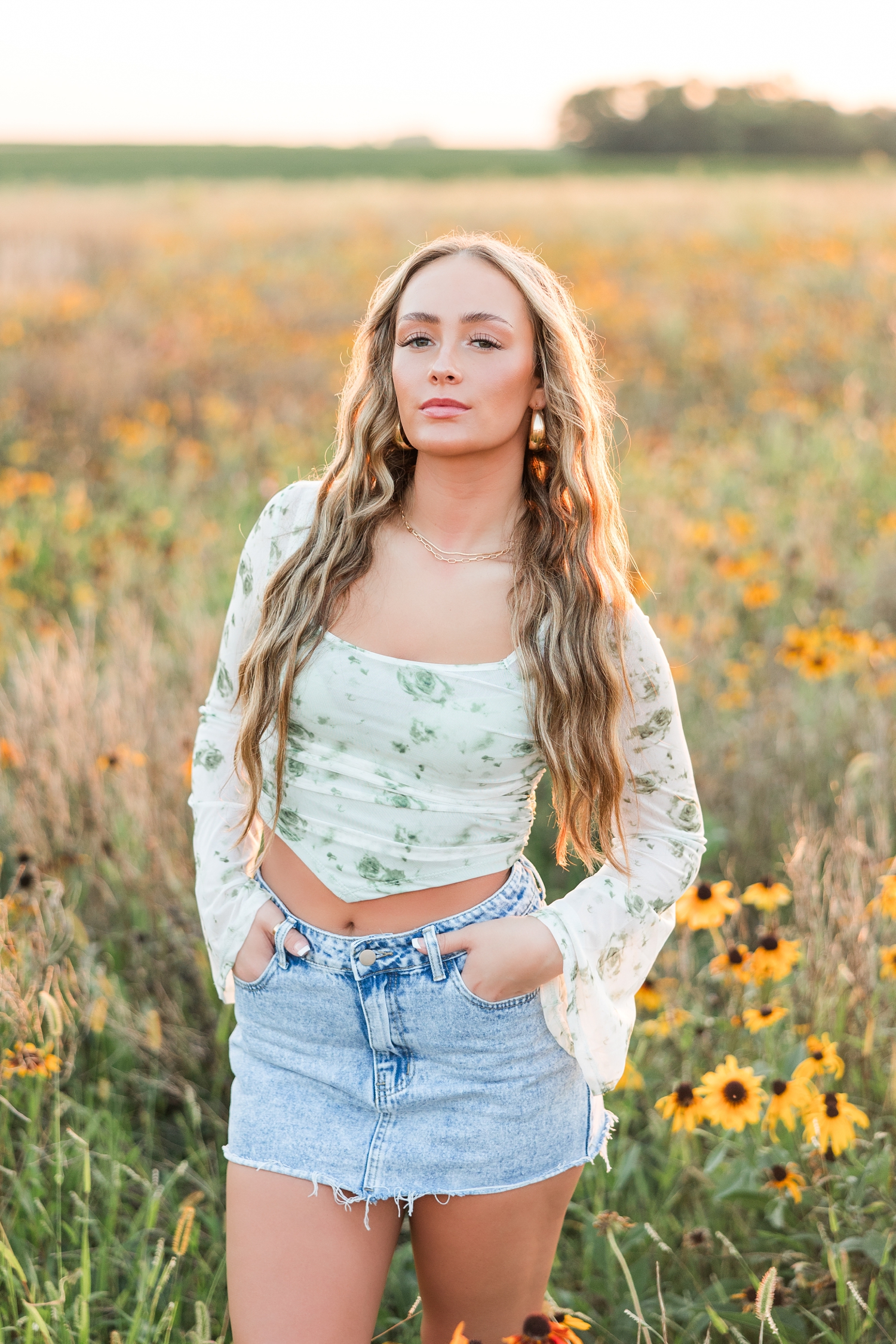 Blaire, wearing a denim mini skirt and green floral long sleeve top, stands with her hands in her pockets in the middle of a wildflower field during golden hour | CB Studio