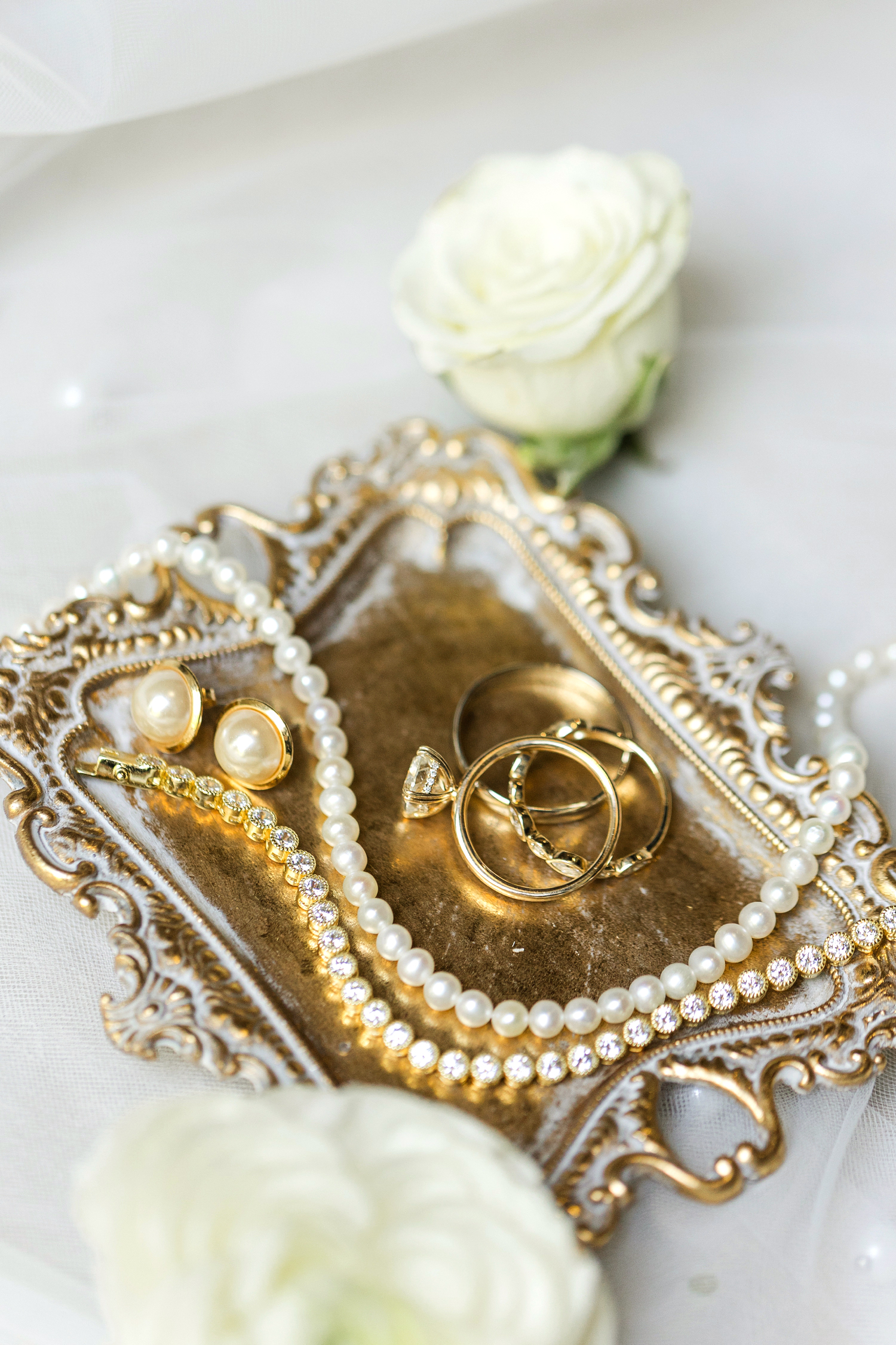 Halie and Nick's wedding bands displayed on a vintage gold tray with Halie's pearl necklace, earrings and diamond tennis bracelet surrounded by white roses | CB Studio