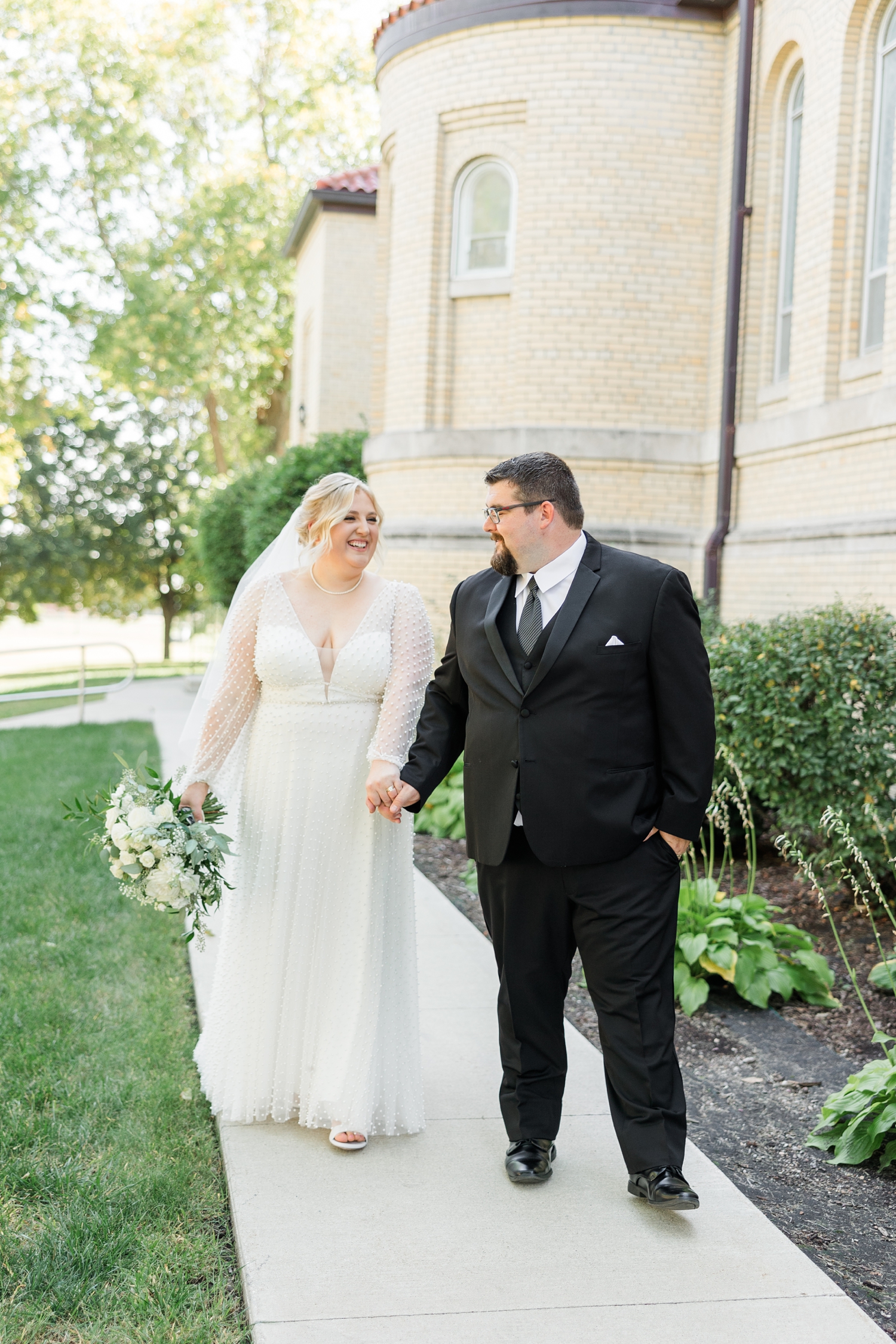 Nick leads his new bride, Halie, down a sidewalk at St. John the Baptist Catholic Church in Bancroft, IA | CB Studio