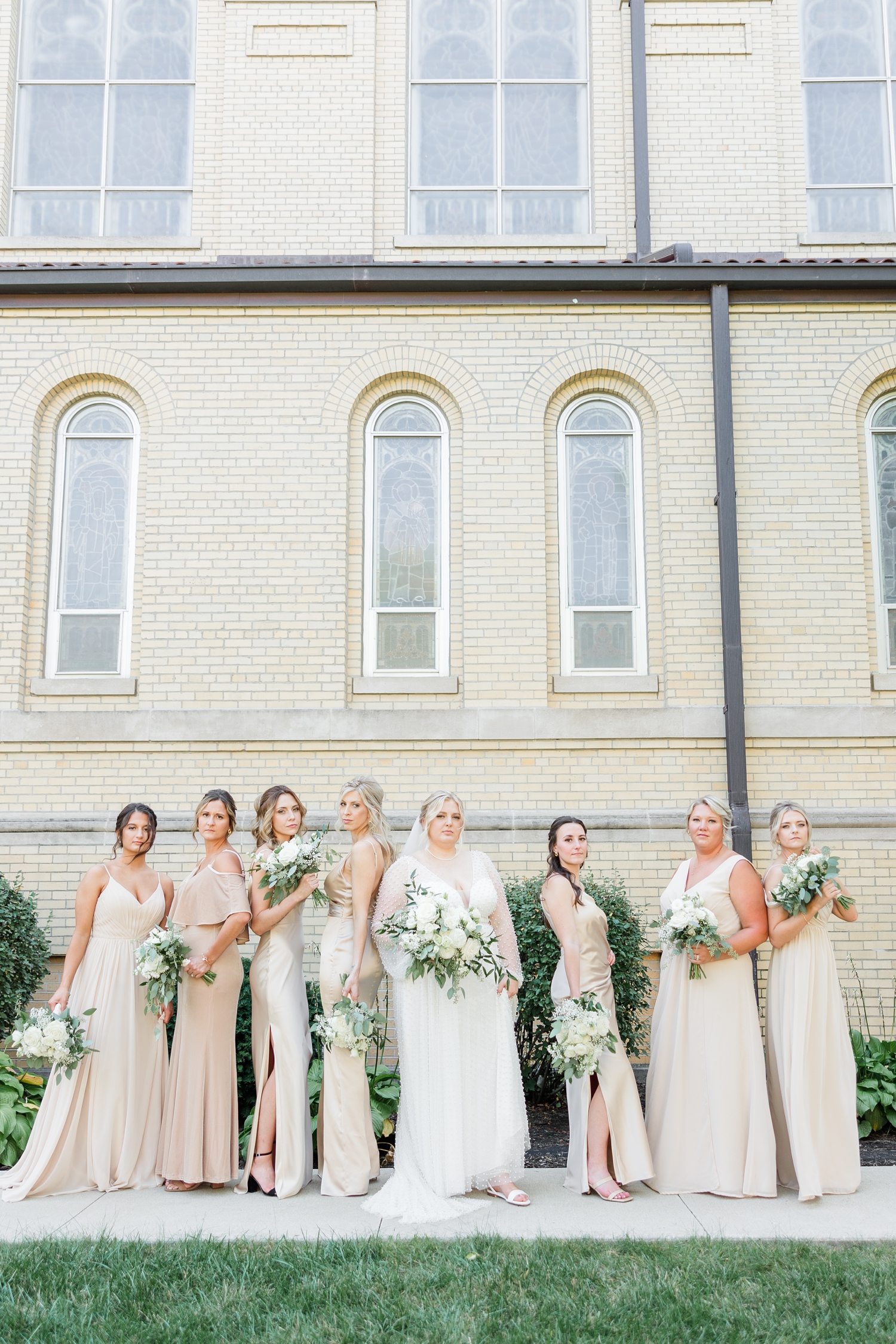 Halie and her bridesmaids, wearing champagne colored dresses, model pose in a line at St. John the Baptist Catholic Church in Bancroft, IA | CB Studio