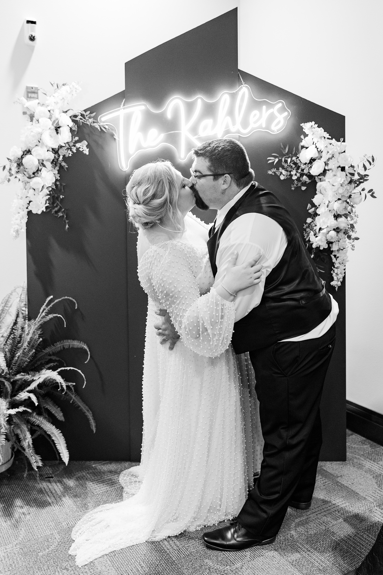 Halie and Nick kiss in front of a backdrop displaying their last name in neon lights surrounded by floral arrangements at The Shores at Five Island | CB Studio