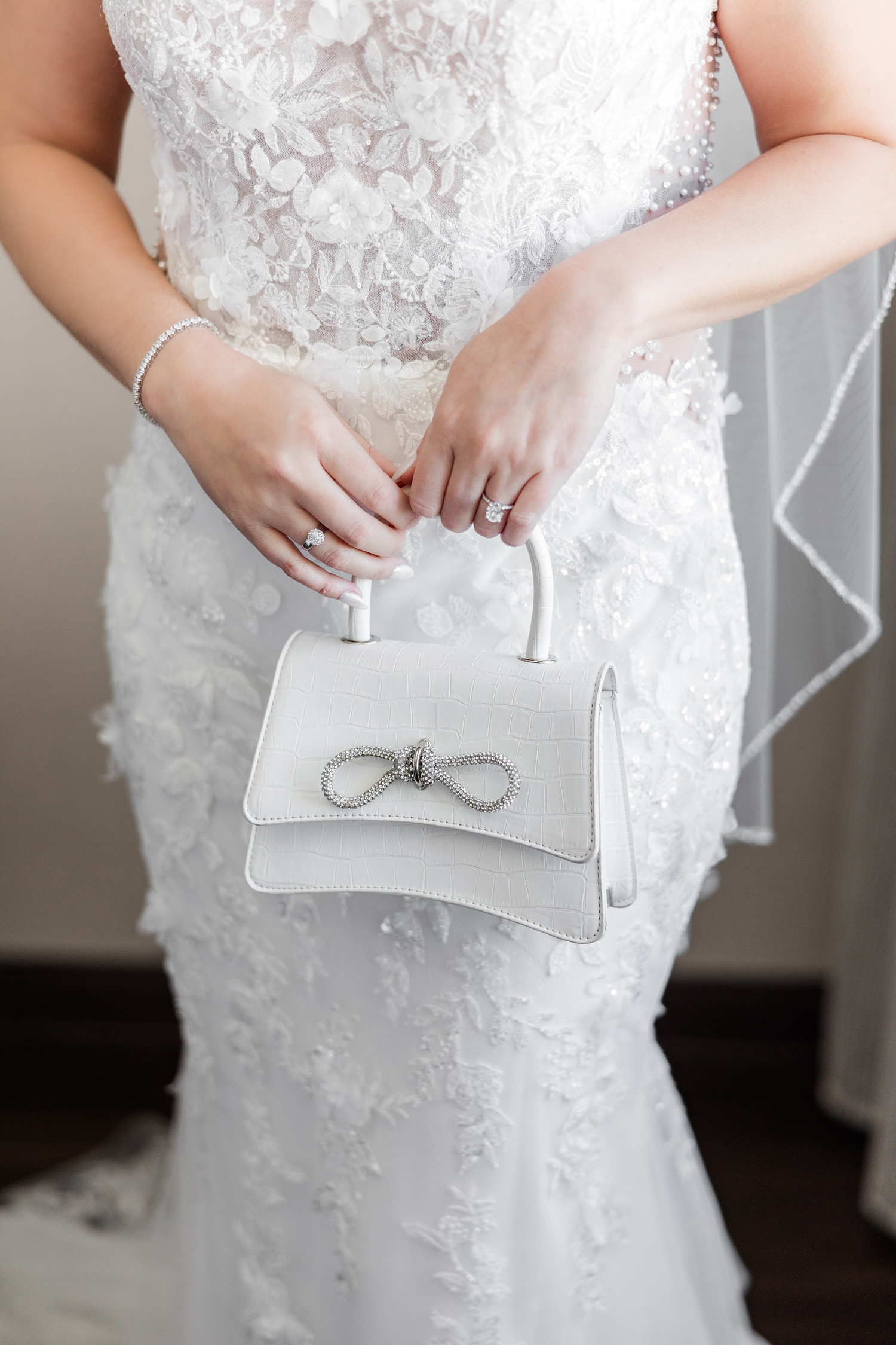 Jenna holds her designer inspired bag with rhinestone encrusted bow in front of her torso while wearing her Morilee 3D detail wedding dress | CB Studio