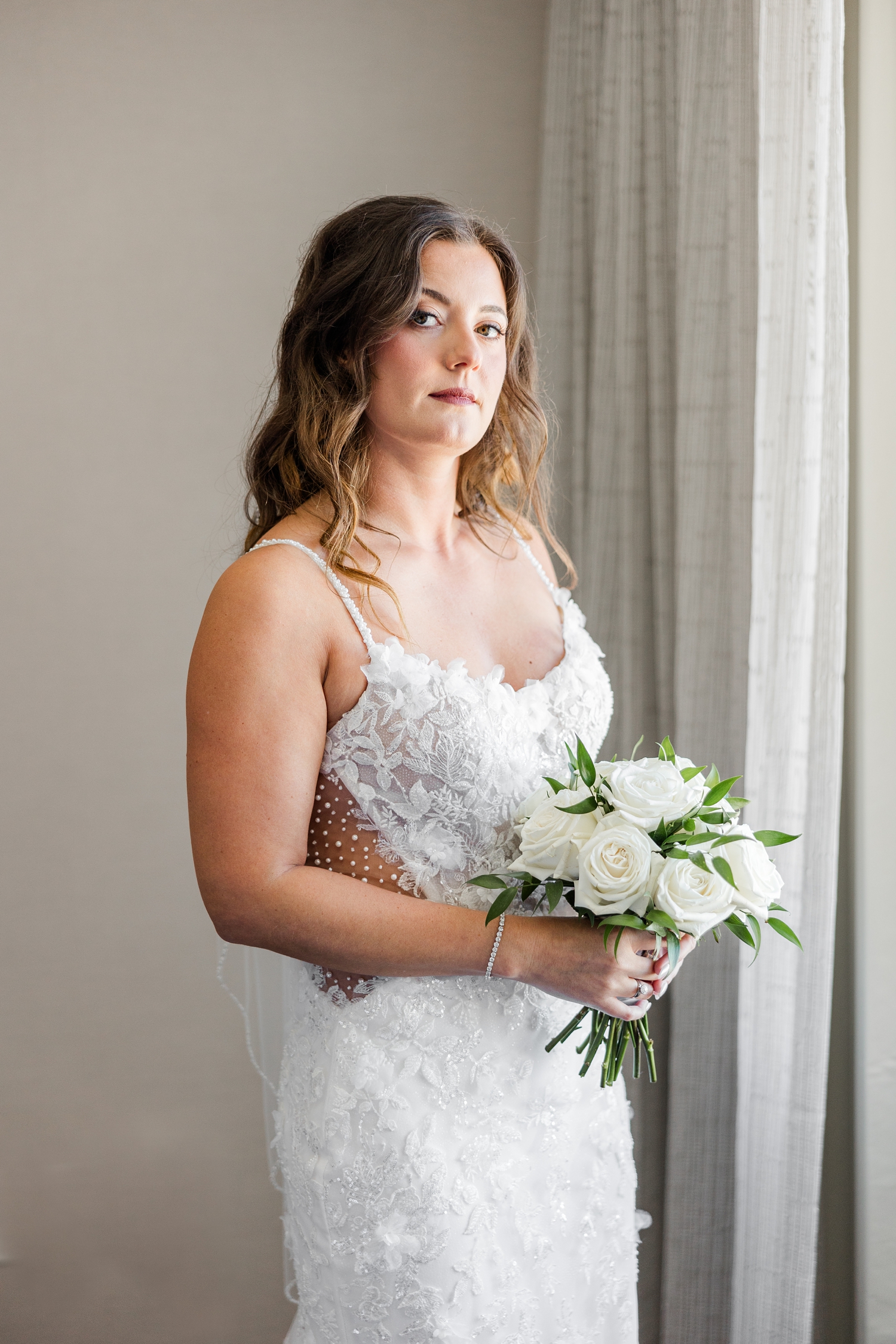 Jenna, standing in her Morilee wedding gown, stares intently while holding her classic white rose wedding bouquet in the bridal suite of the Downtown Des Moines Marriott | CB Studio