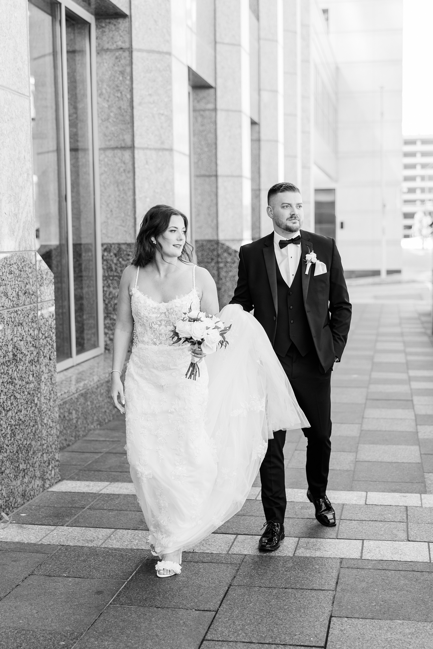 Dustin holds Jenna's wedding dress train while they look to the right as they walk across the back entrance of 801 Grand in downtown Des Moines | CB Studio