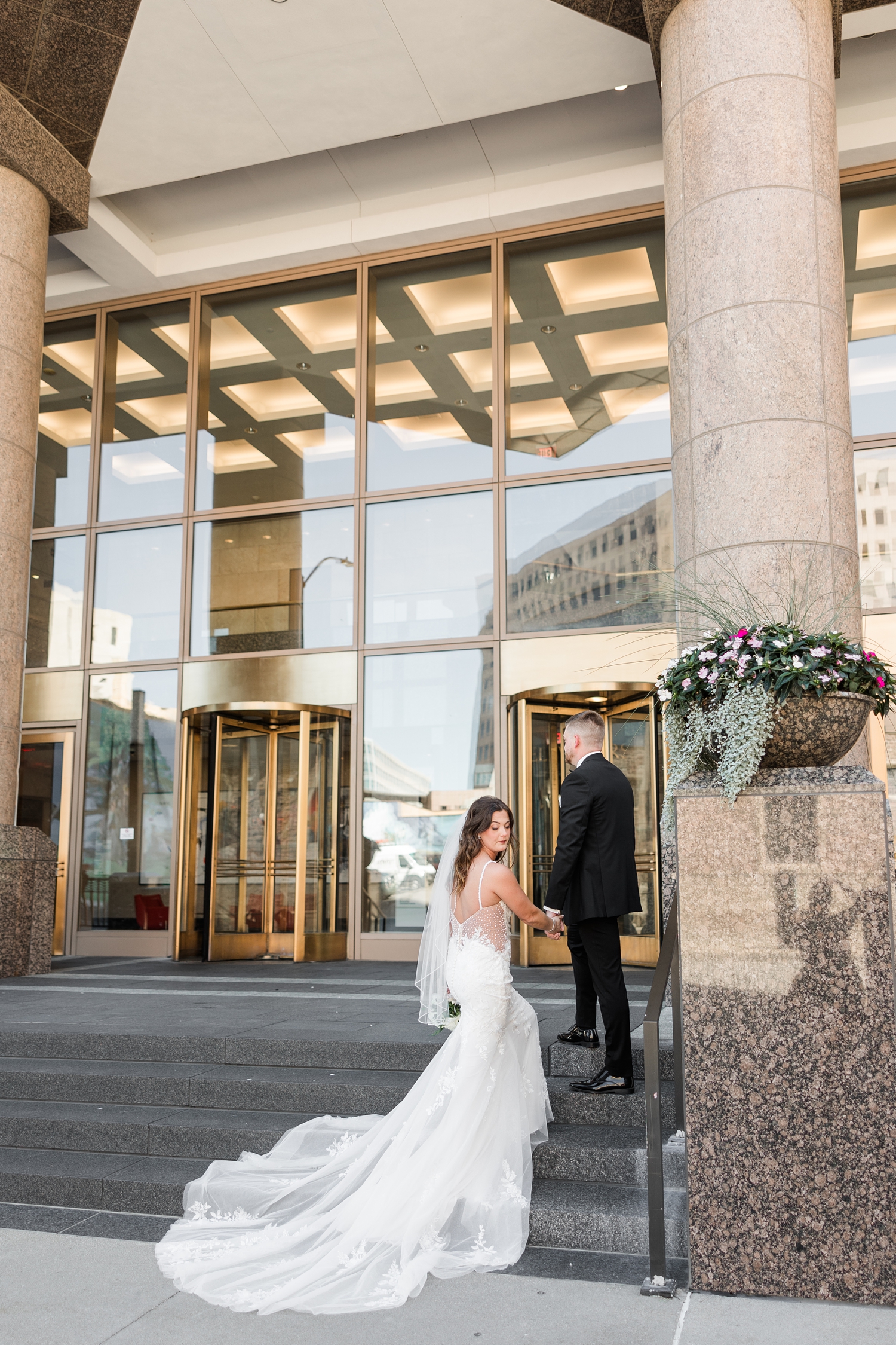 Dustin and Jenna walk up the steps of 801 Grand in downtown Des Moines while looks back at her wedding dress train | CB Studio